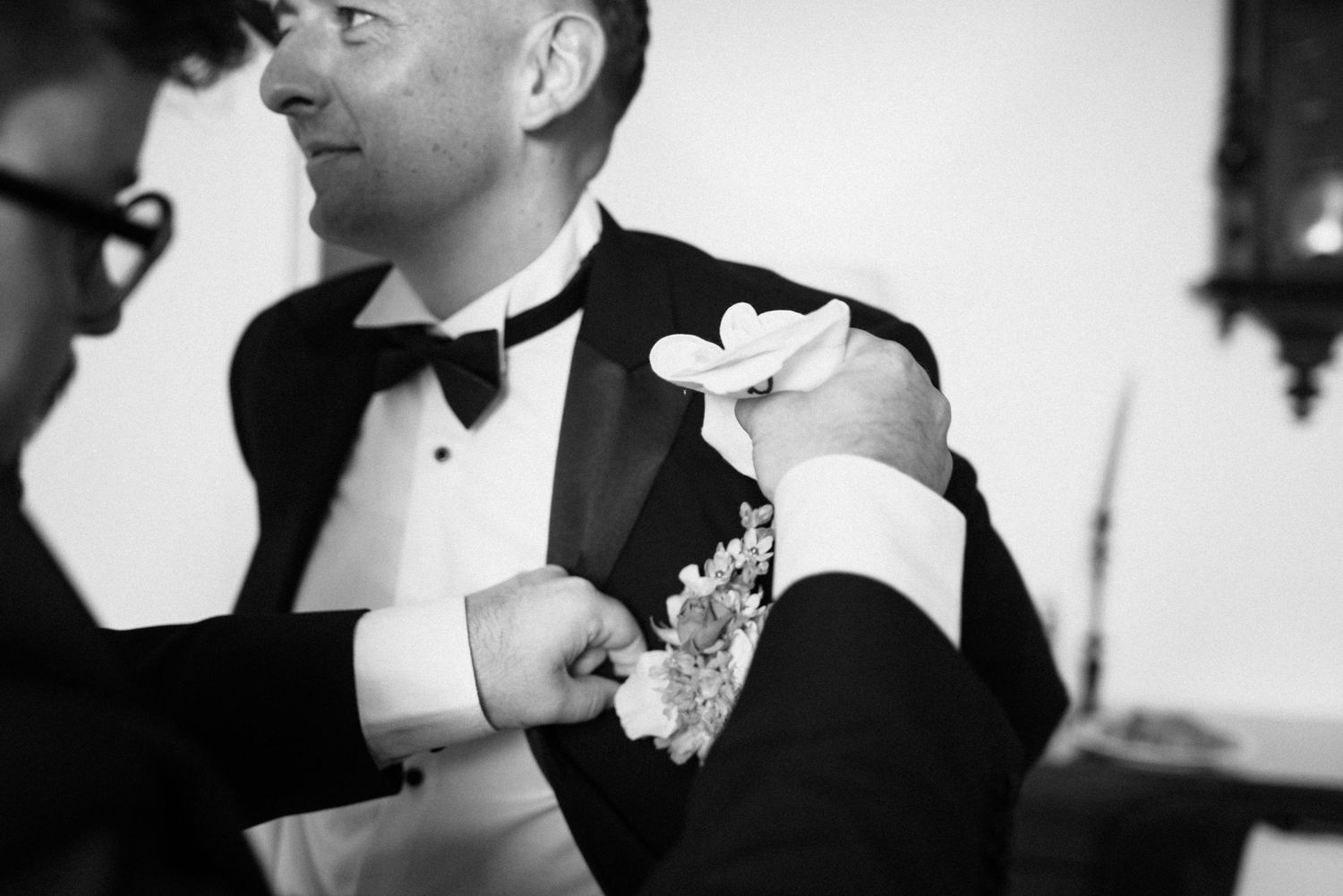 Black and white photo of a groom having his boutonniere pinned on his tuxedo lapel at a wedding.