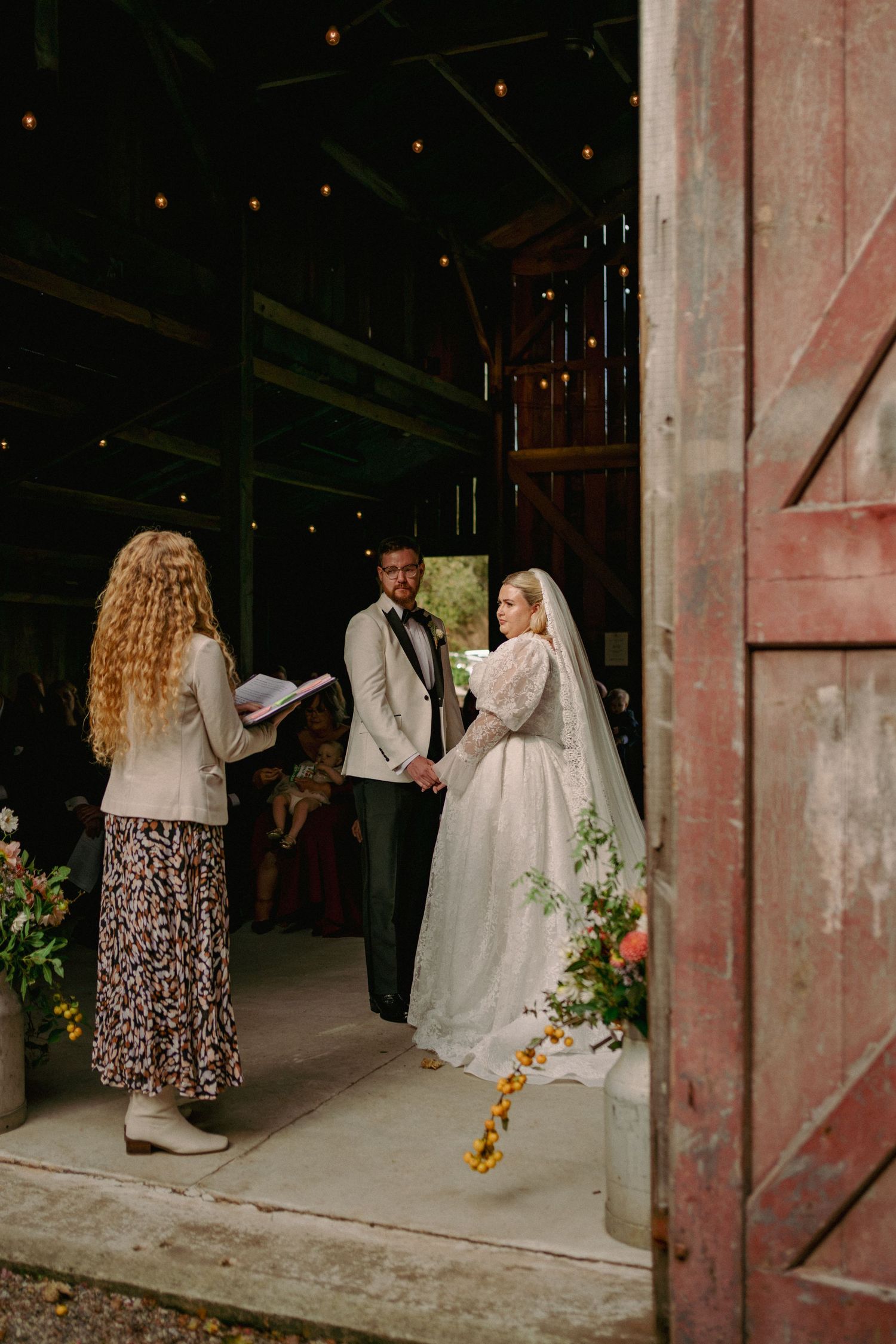 Bride and groom hold hands during a rustic barn wedding ceremony as an officiant reads, with string lights and floral arrangements nearby.