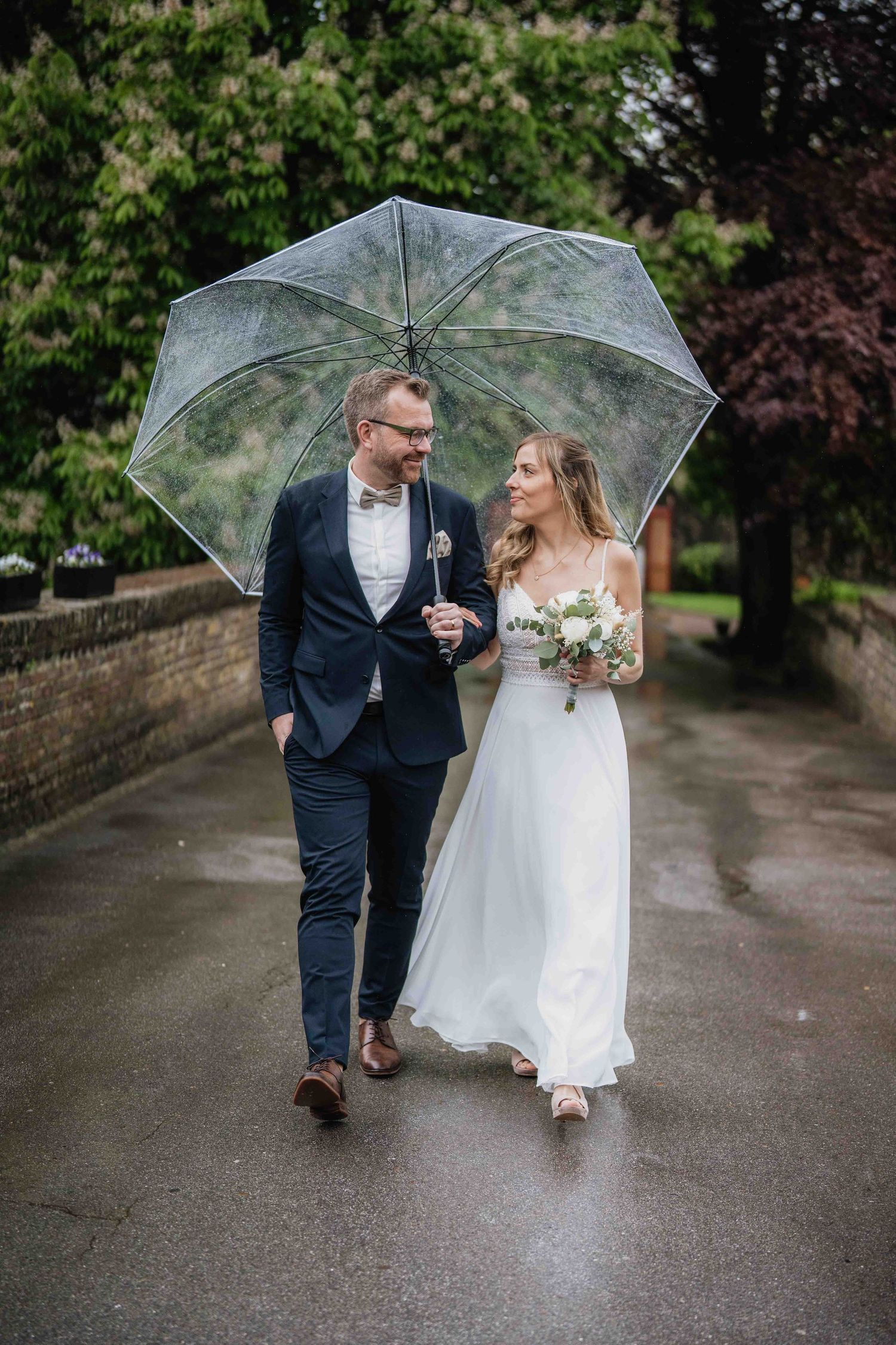 A couple walking together under a clear umbrella on a rainy day path surrounded by lush greenery.