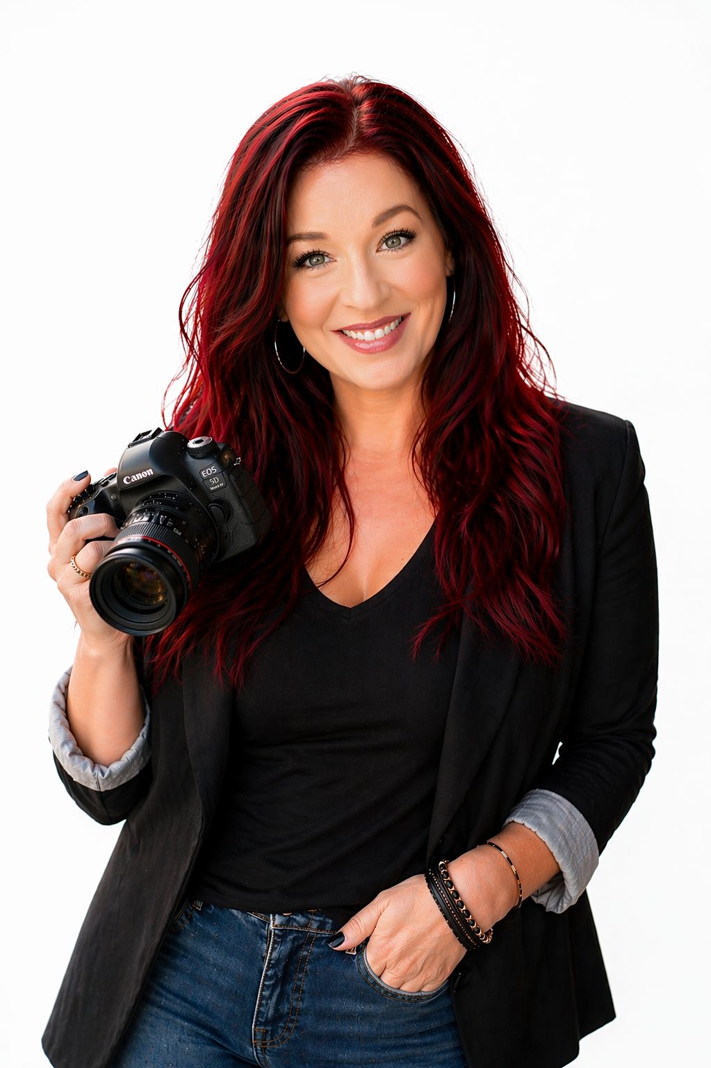 Woman with red hair holding a DSLR camera, wearing a black blazer and jeans, smiling against white background.