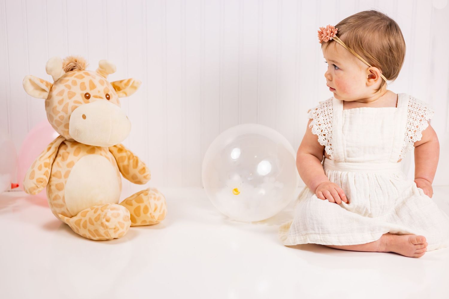 Baby in white dress sitting beside plush giraffe toy on white background with transparent balloon.