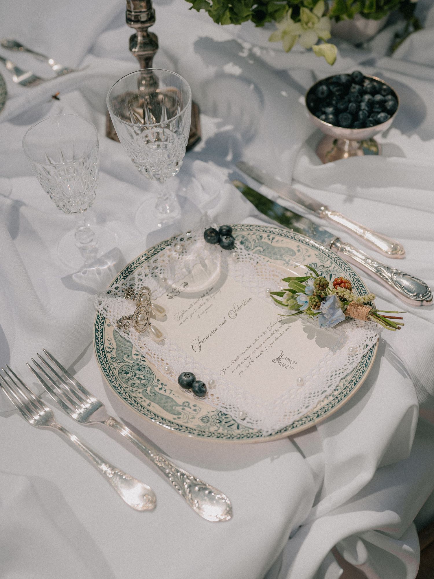 Elegant table setting with crystal glassware, ornate plate, and blueberries on white linen tablecloth with silver cutlery.