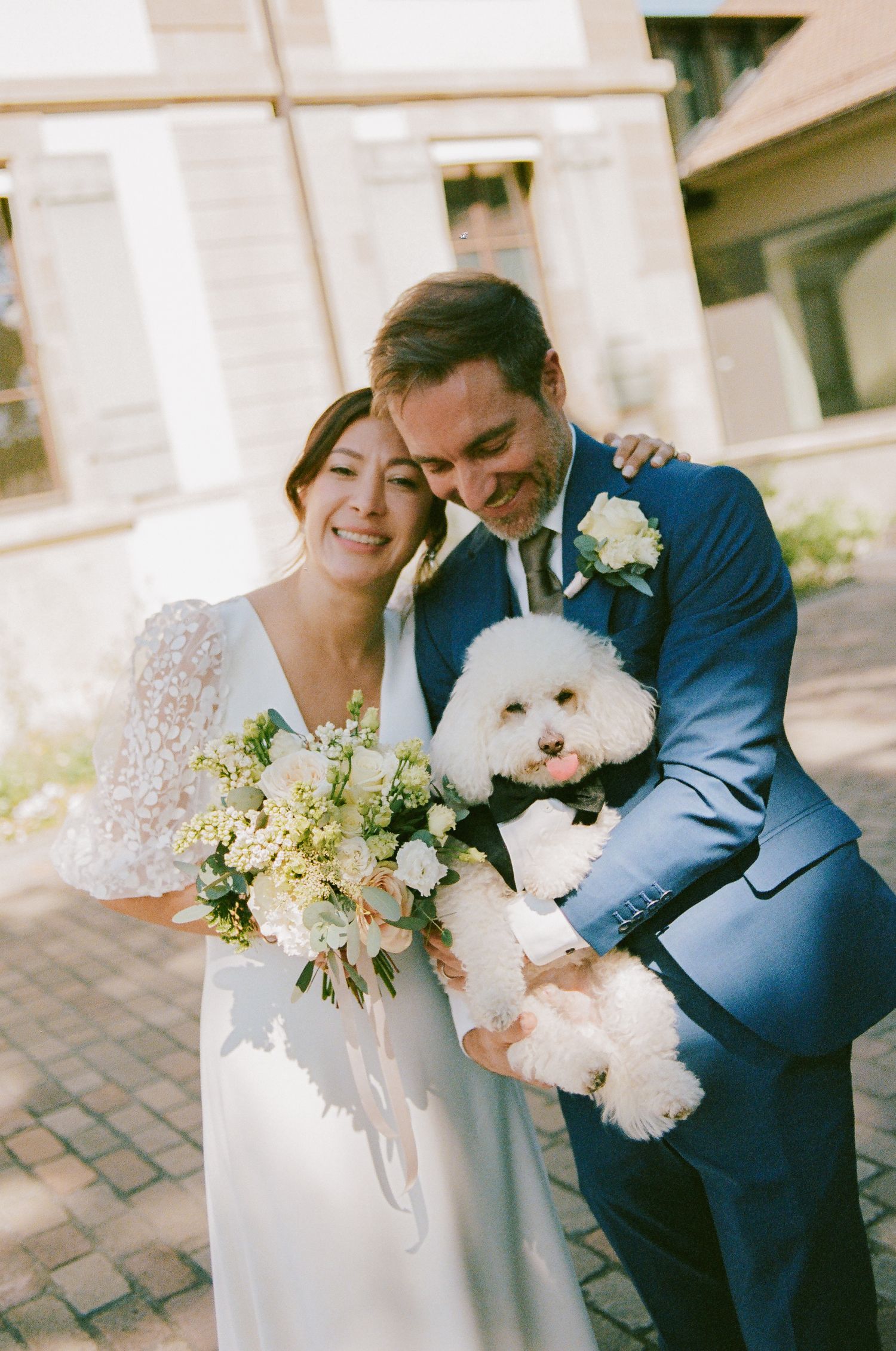 Smiling bride and groom in blue suit holding white Bichon Frise dog, bride carries white floral bouquet at outdoor wedding.