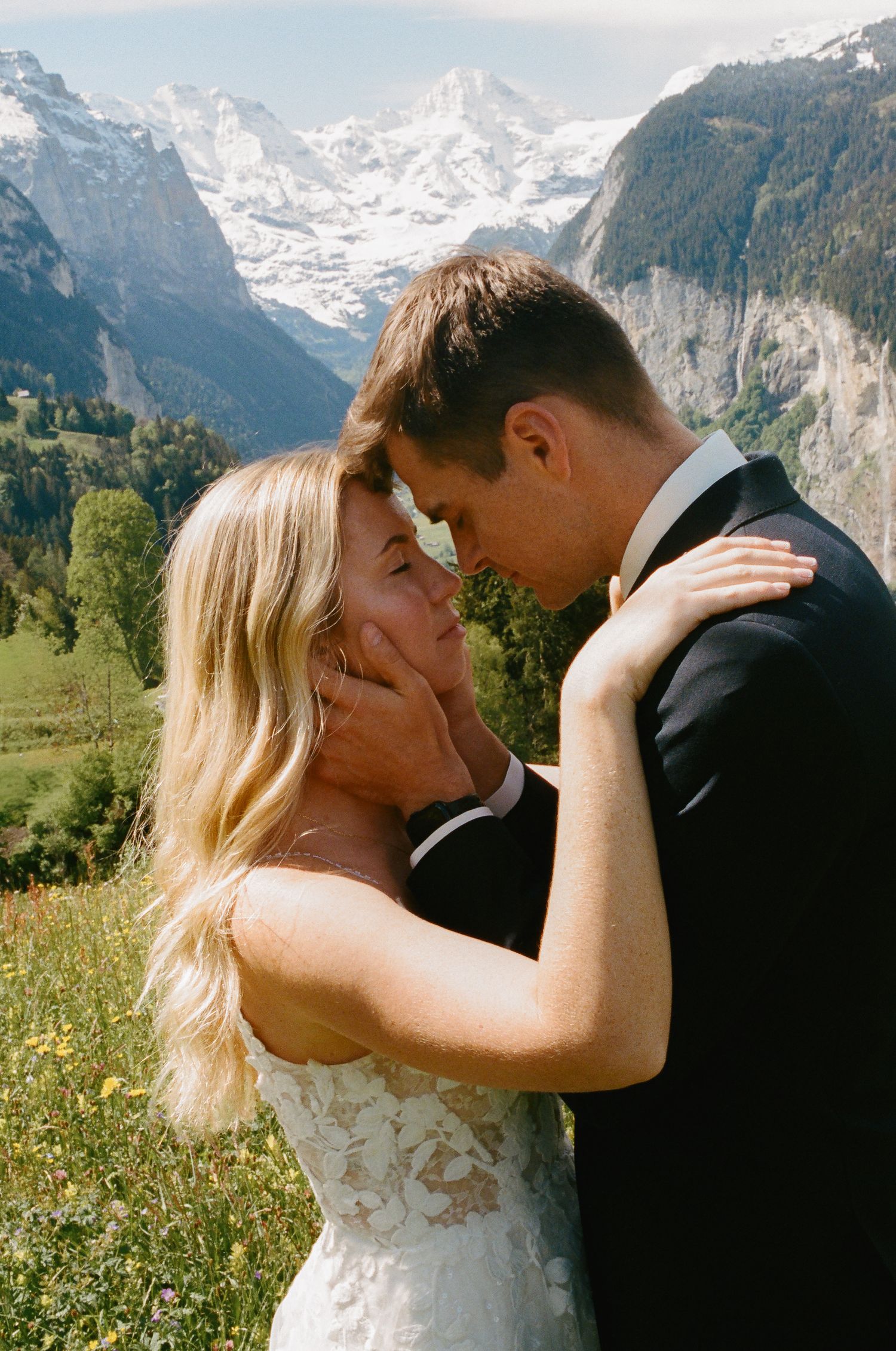 A couple in wedding attire shares a romantic moment with Swiss Alps mountains and wildflowers in the background.