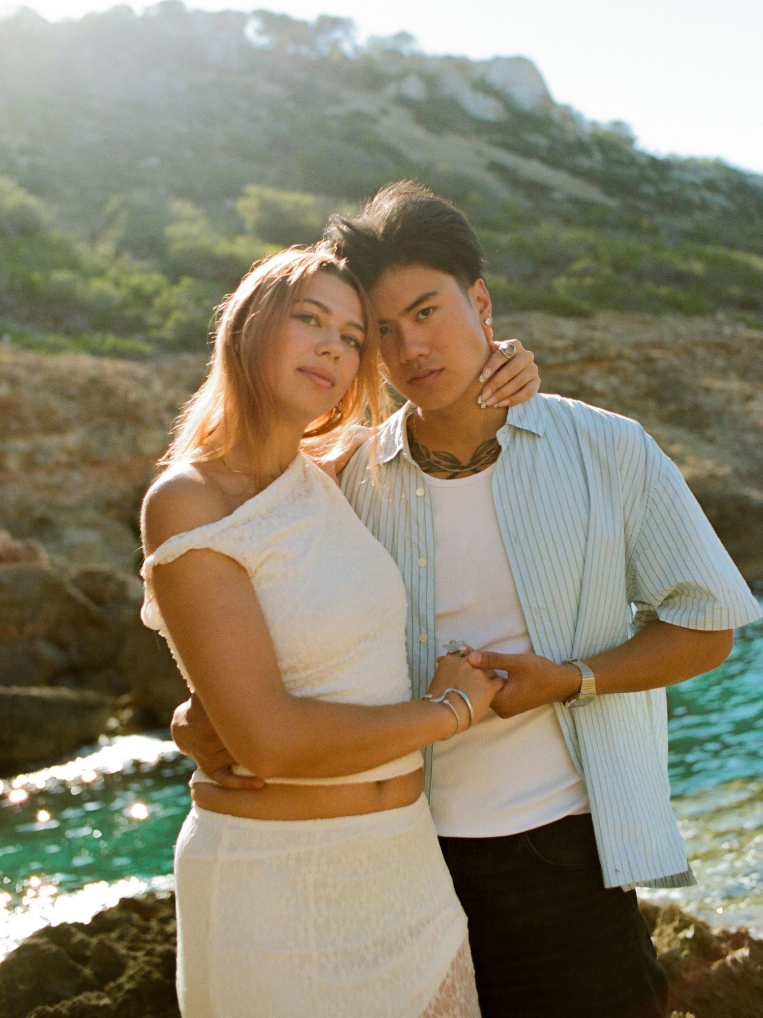 A couple poses together on rocky coastline with sunlit ocean and mountains in background during golden hour.