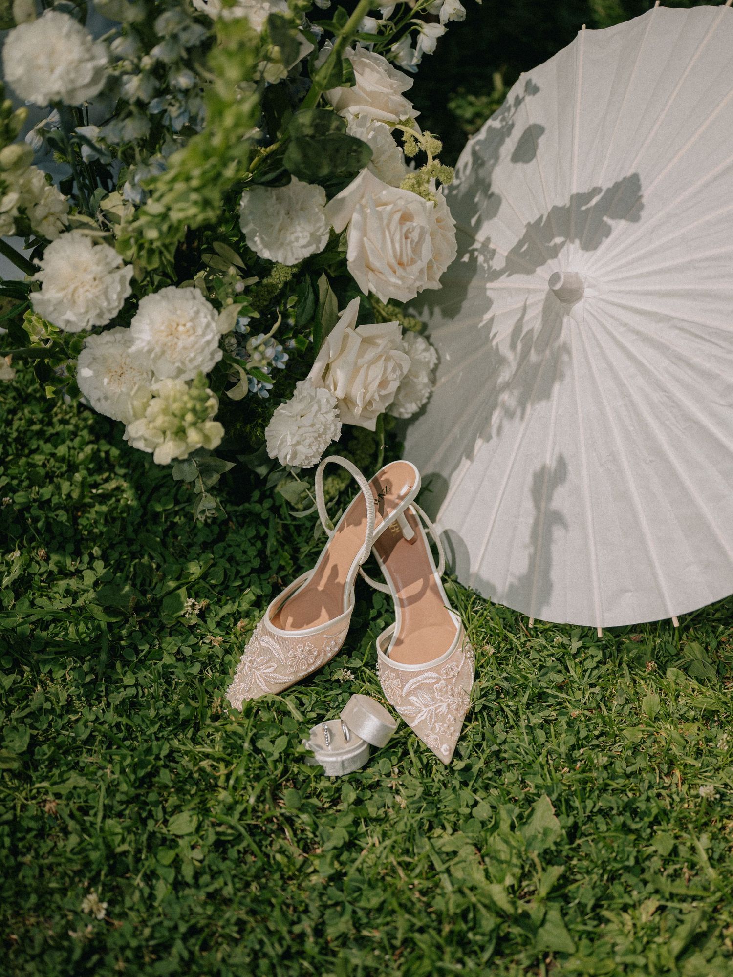 Delicate lace wedding shoes arranged on grass with white parasols and floral decorations in an outdoor setting.
