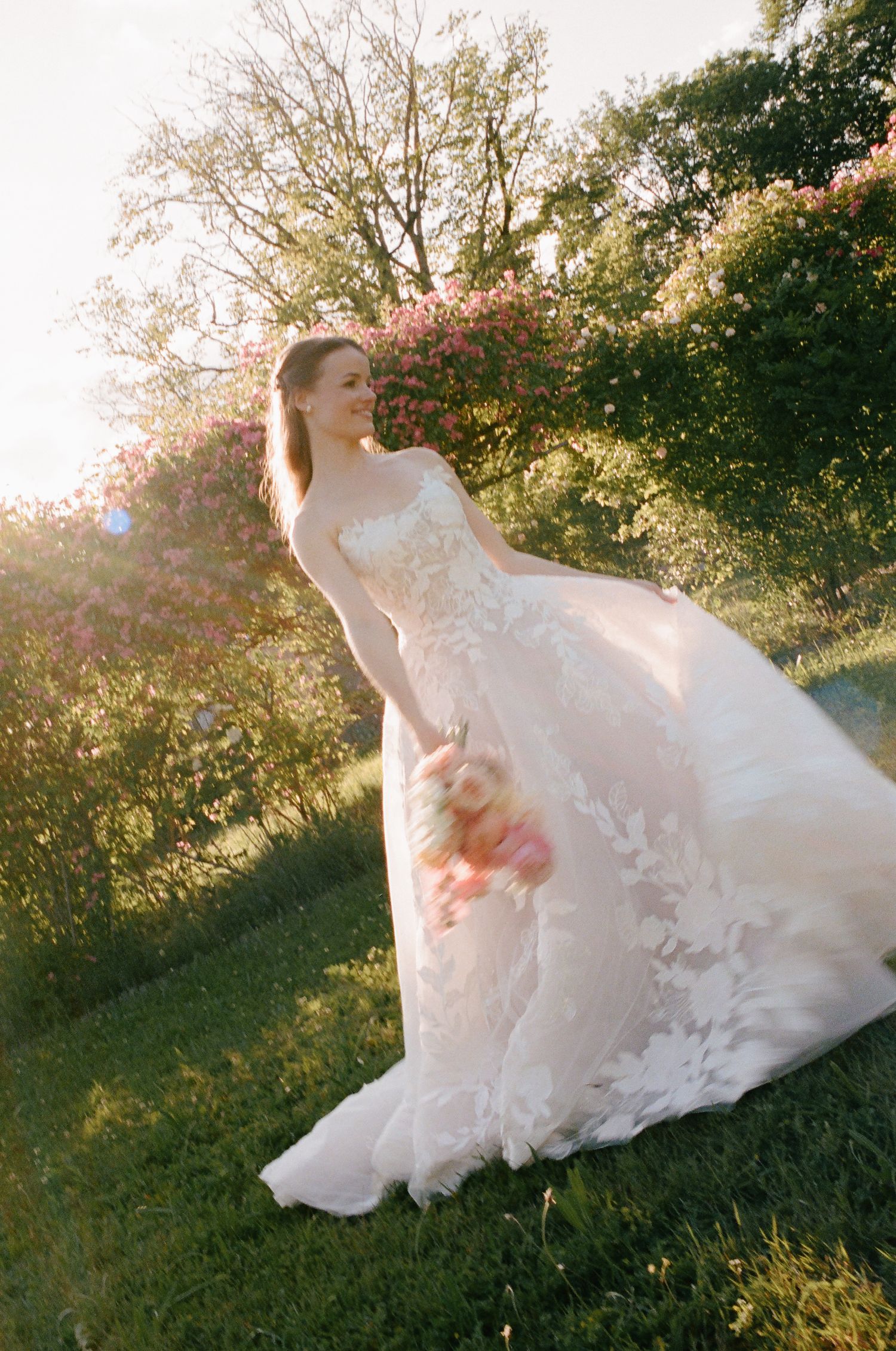 Bride in floral lace wedding gown holding bouquet in sunlit garden with blooming pink roses and green trees.