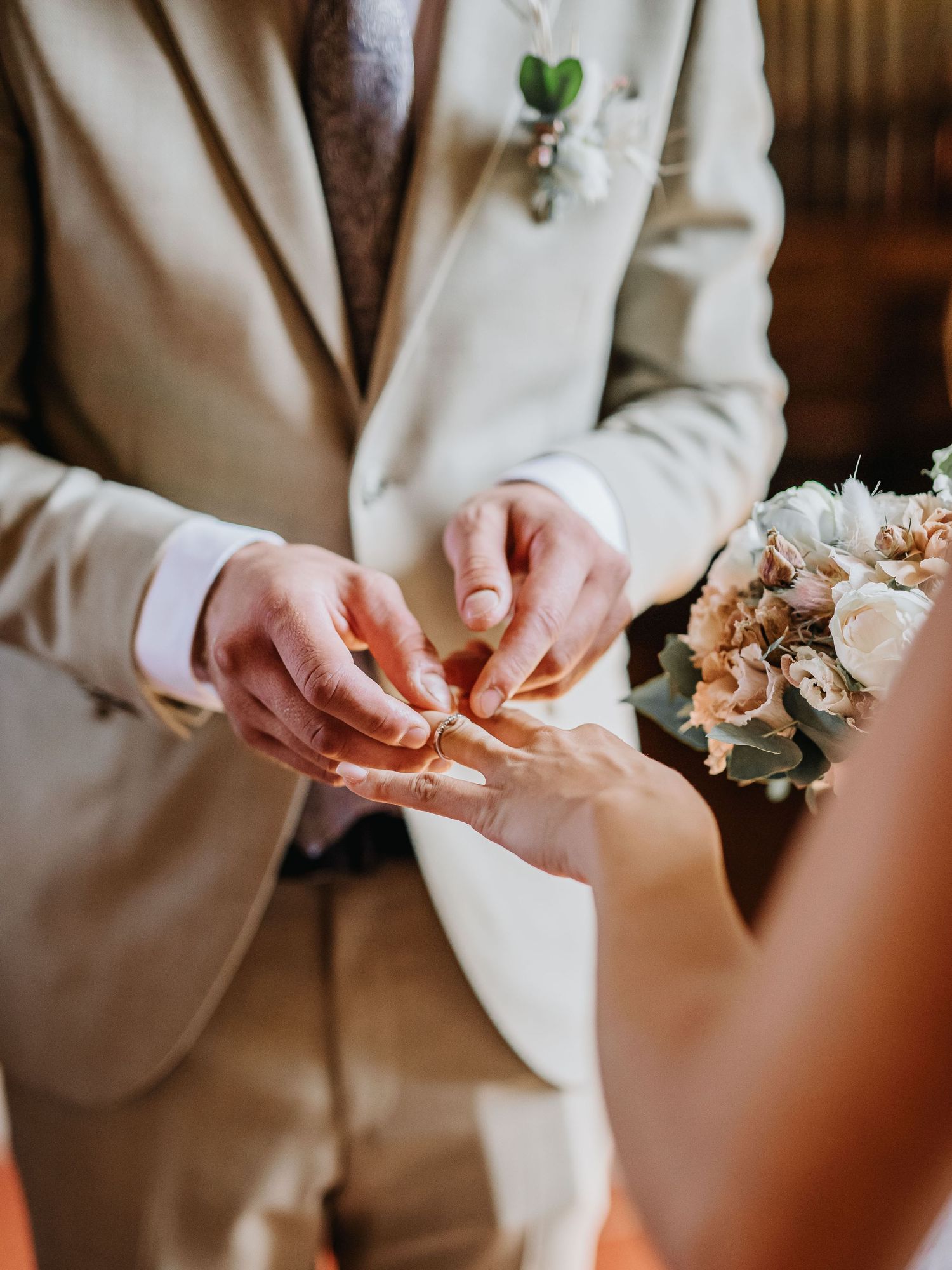 Close-up of hands exchanging wedding rings during ceremony with peach bouquet visible.