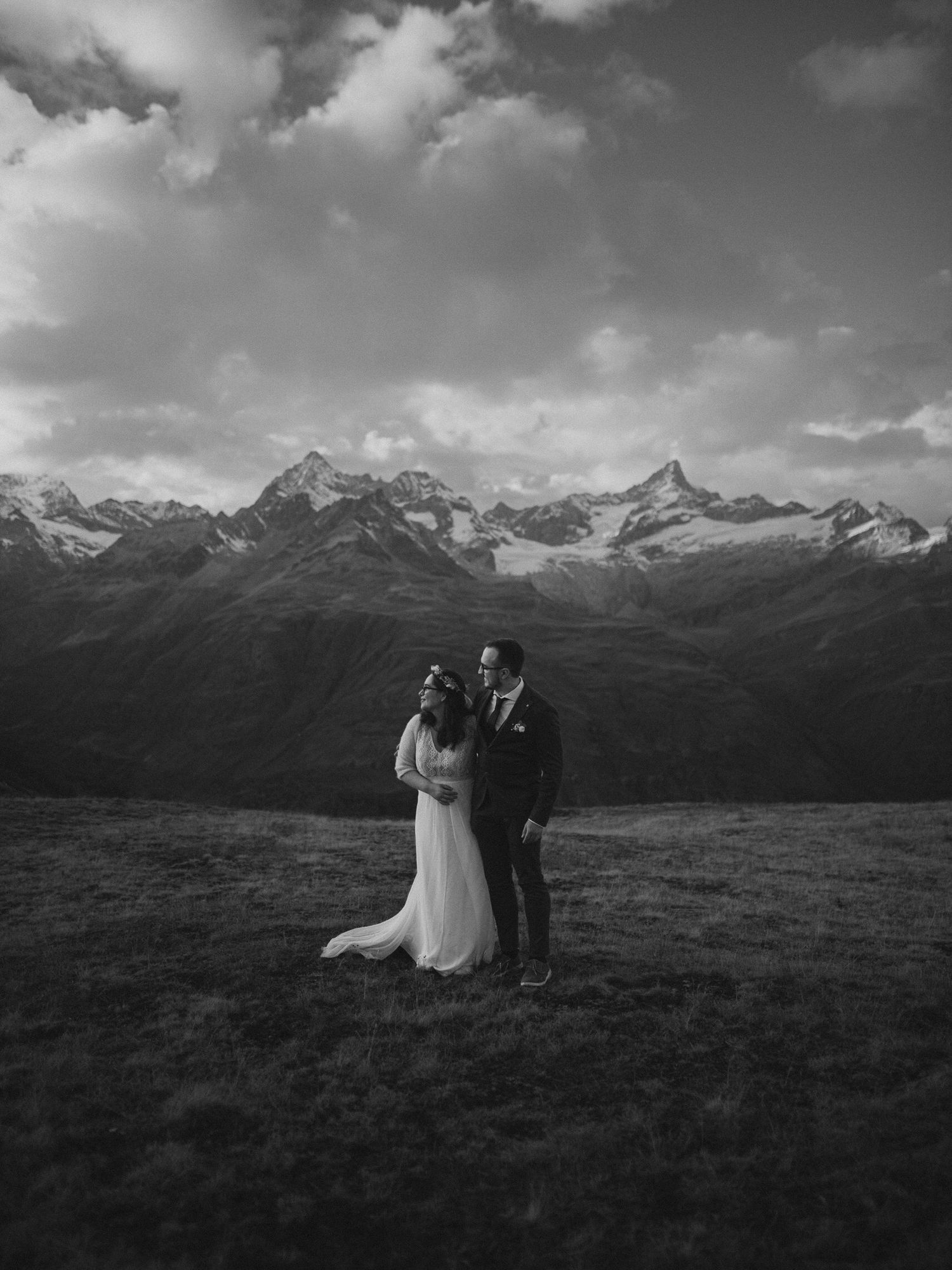 A panoramic black and white wedding scene unfolds against dramatic mountain peaks and cloudy skies in a vast meadow.