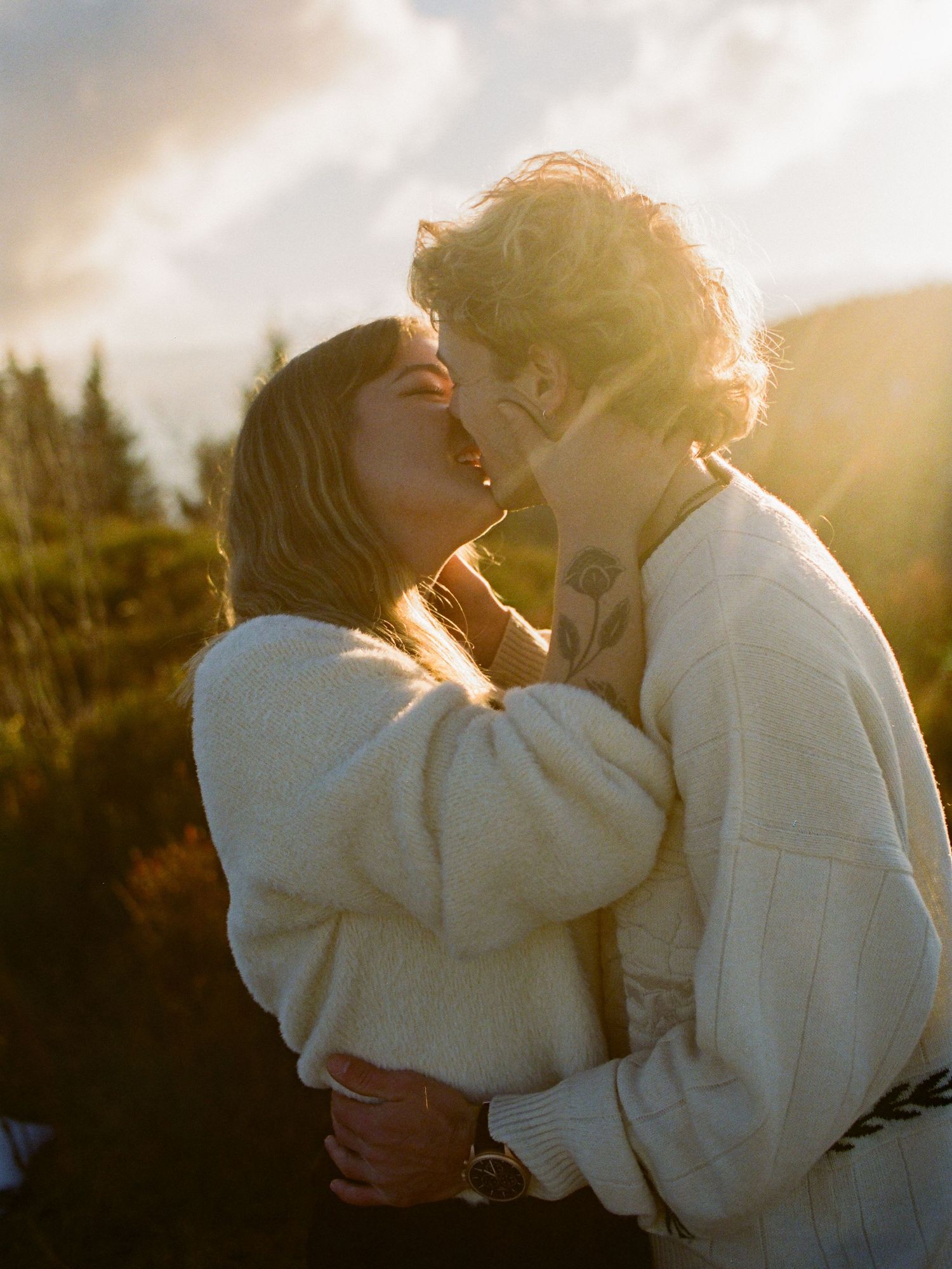 Couple sharing a romantic kiss at golden hour with soft sunlight and mountain forest backdrop.