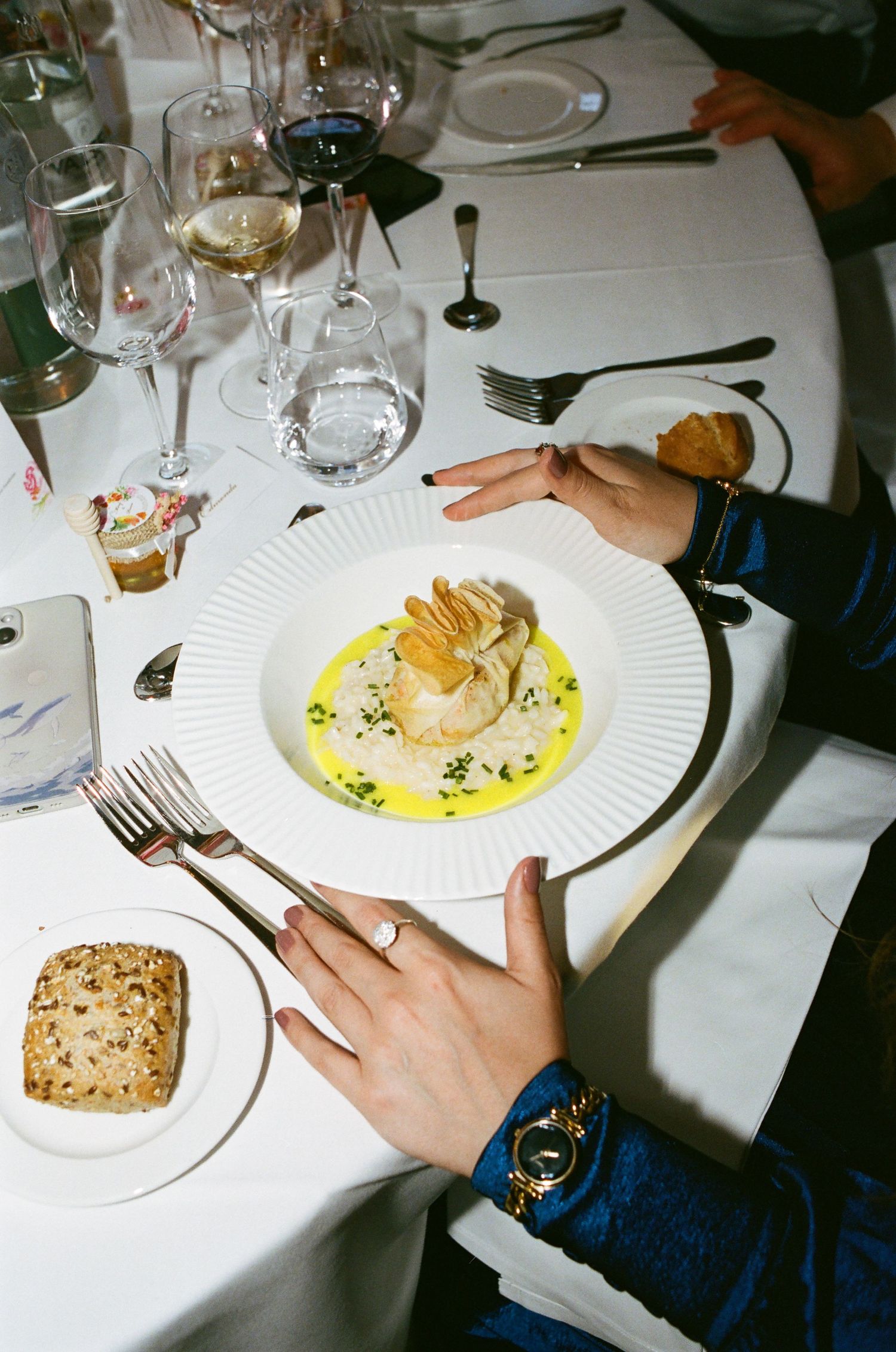 Elegant fine dining plate with scallop in yellow sauce, served on white tablecloth with wine glasses and bread roll.