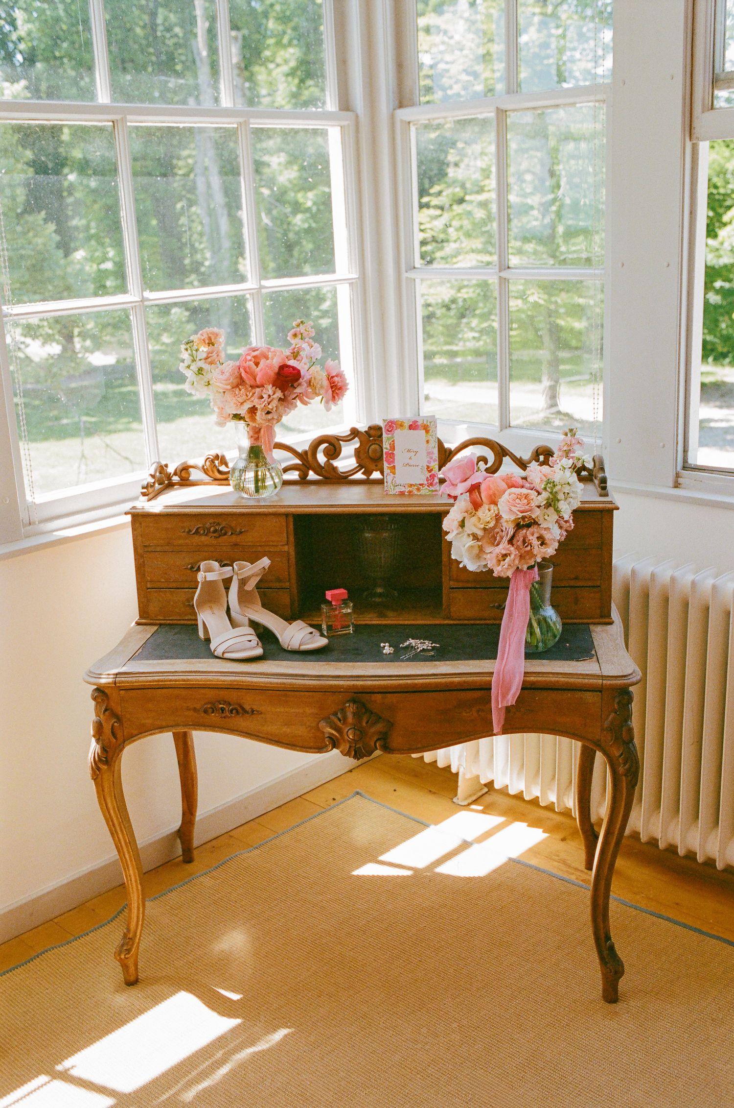 Vintage French writing desk with pink floral arrangements, white heels, and wedding details by window.