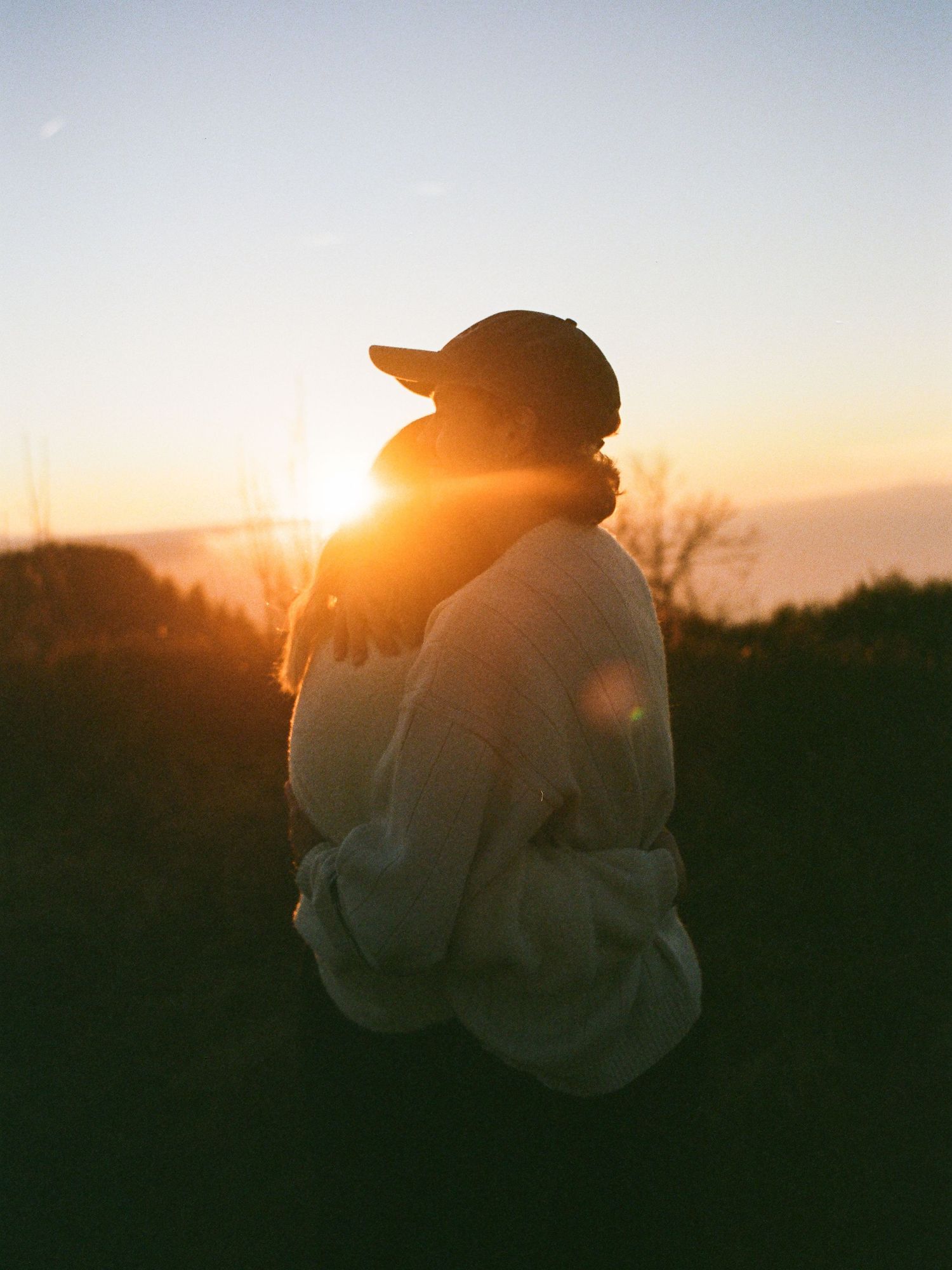 Silhouette of person wearing baseball cap and sweatshirt sits outdoors against dramatic golden sunset sky.