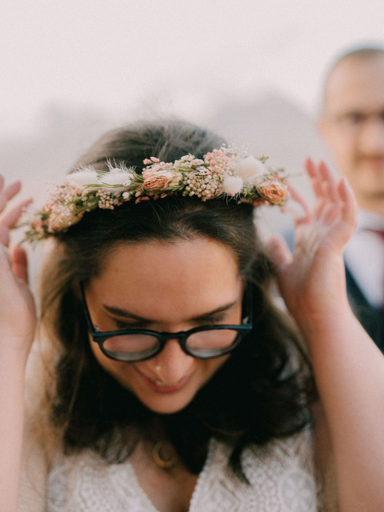 A person wearing glasses and a delicate floral crown made of small dried flowers against a light background.