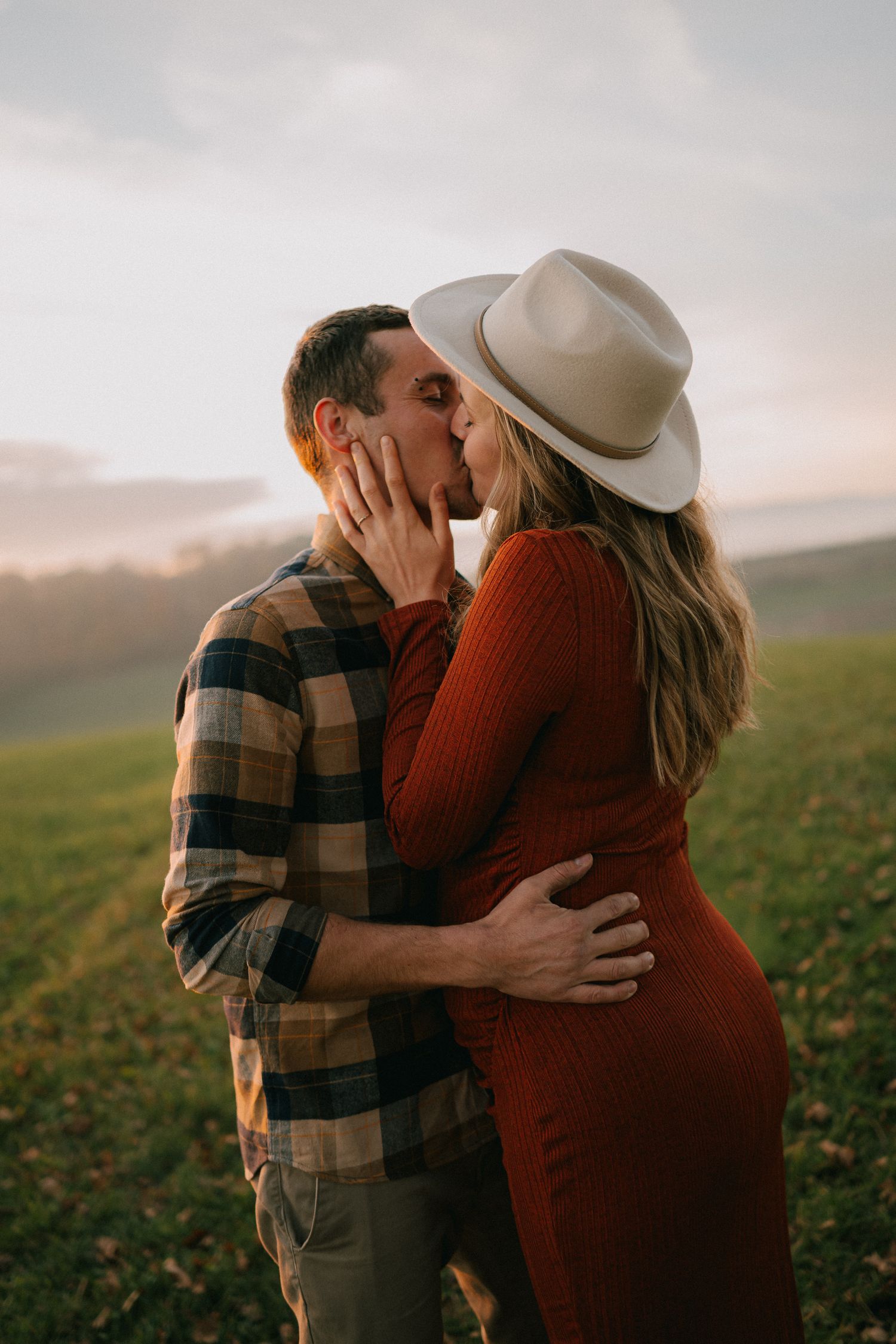 Romantic couple embraces in a field at sunset, with a woman in a red sweater and white hat kissing her partner.