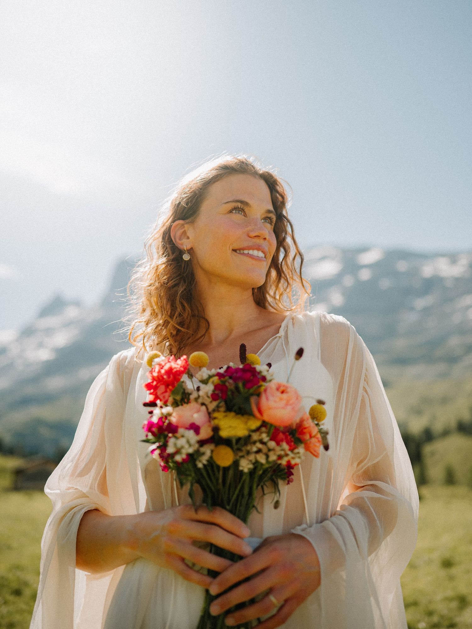 Person in white holding a vibrant bouquet of flowers against a mountain backdrop.