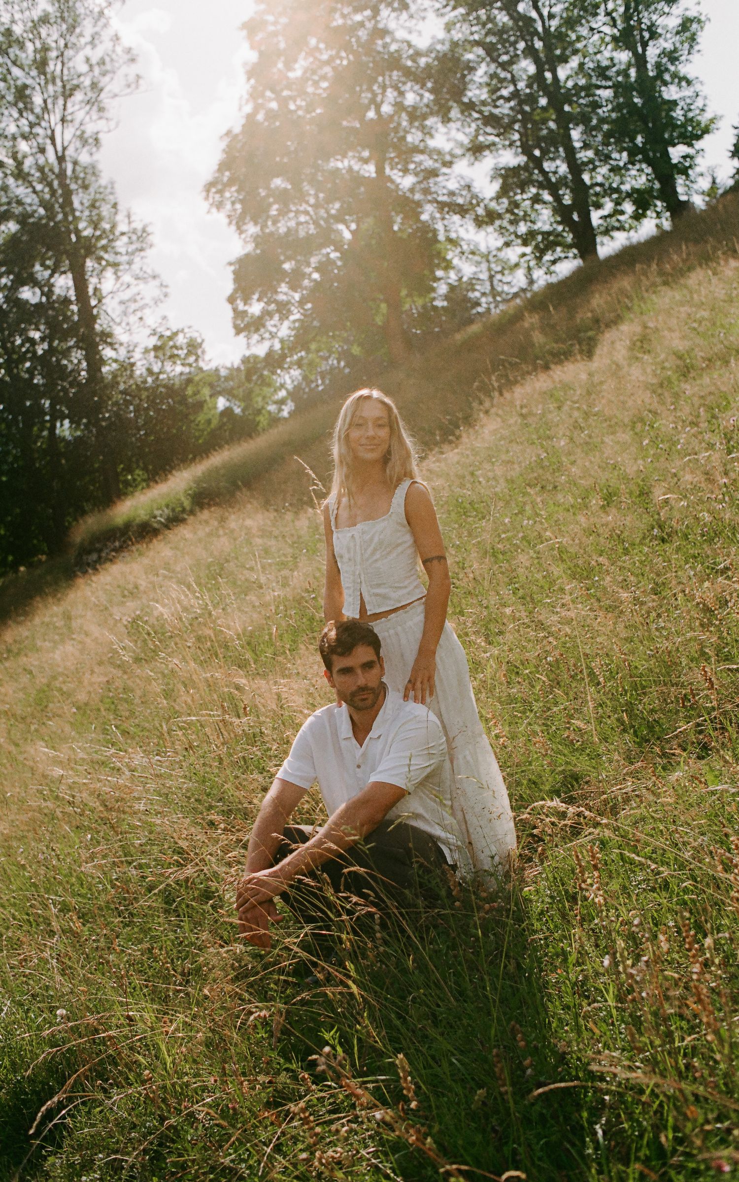 Couple in white outfits posing in a sunlit grassy meadow with trees, romantic outdoor portrait photography.