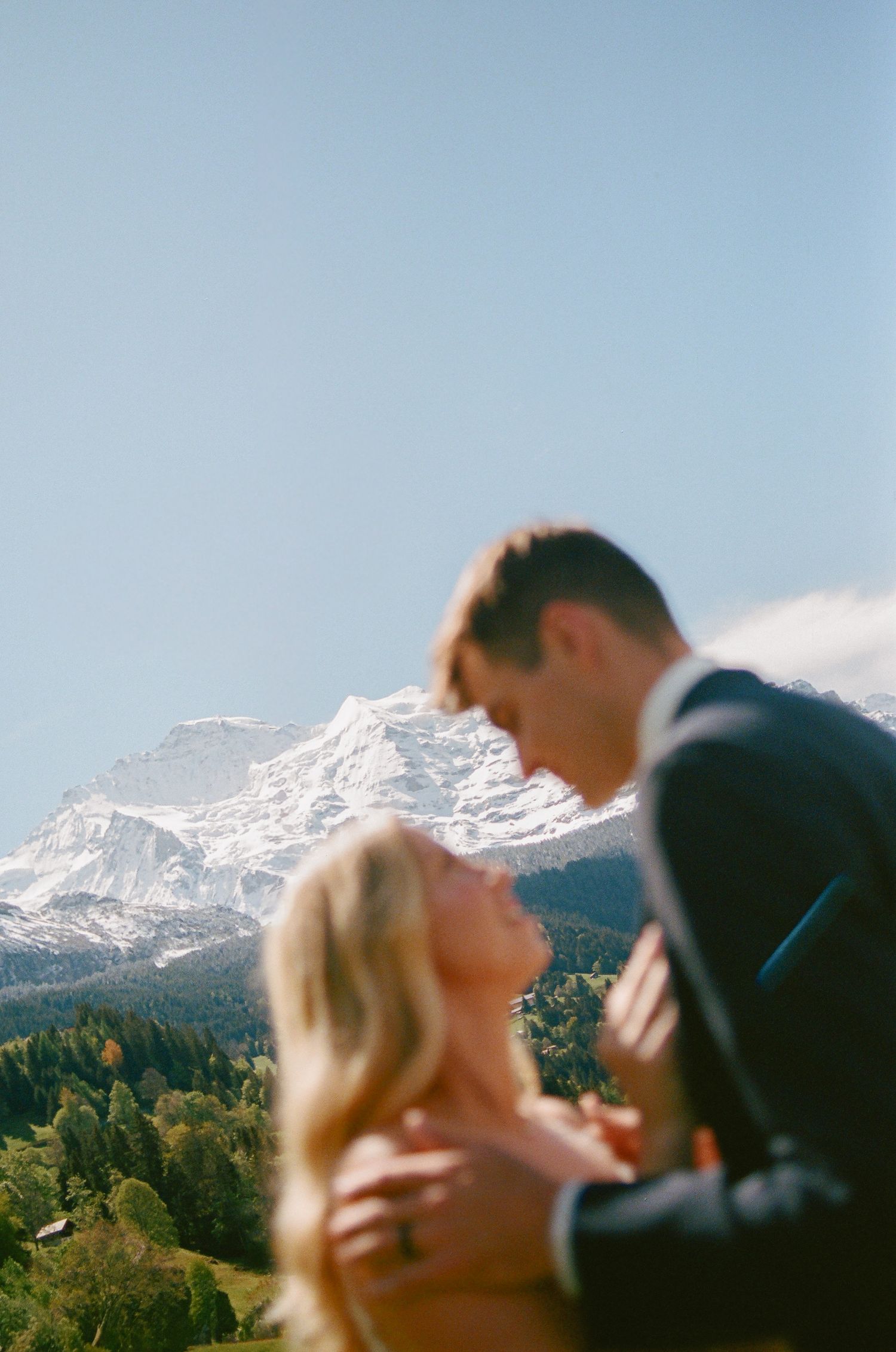 Couple embracing with snow-capped Alps and green forest backdrop on a clear sunny day.