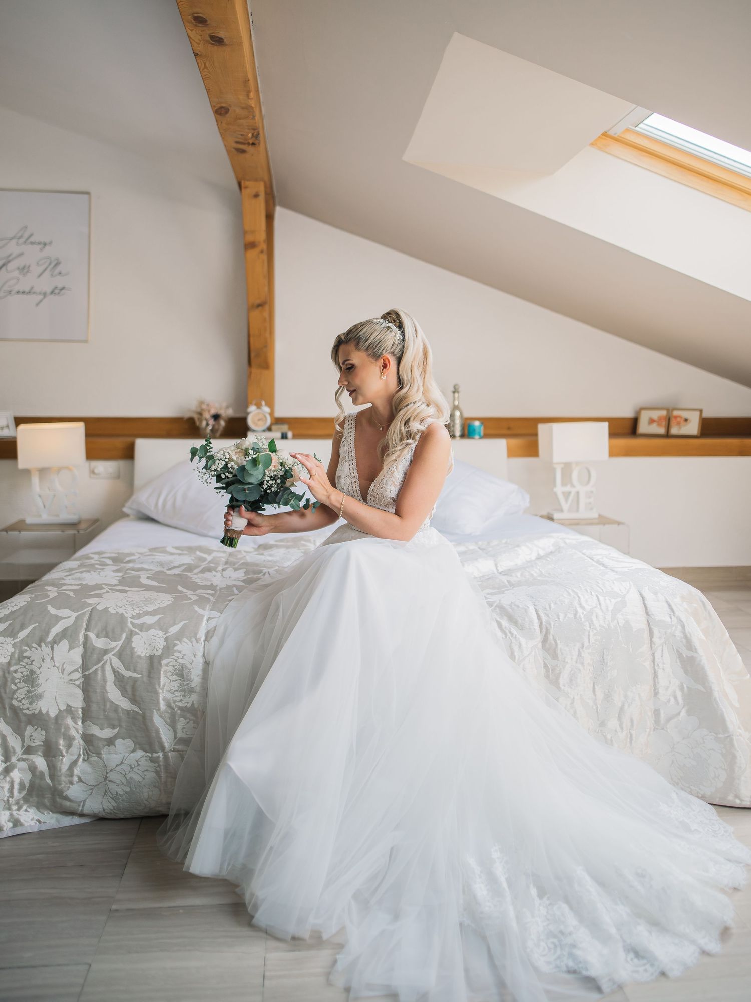 Bride in flowing tulle gown sits on white bedding in rustic attic room with wooden beams.