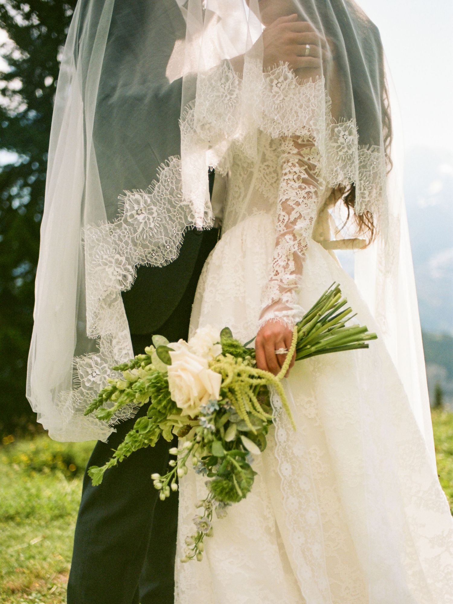 A wedding couple stands on a grassy hilltop with white lace veil and bouquet flowing in the mountain breeze.