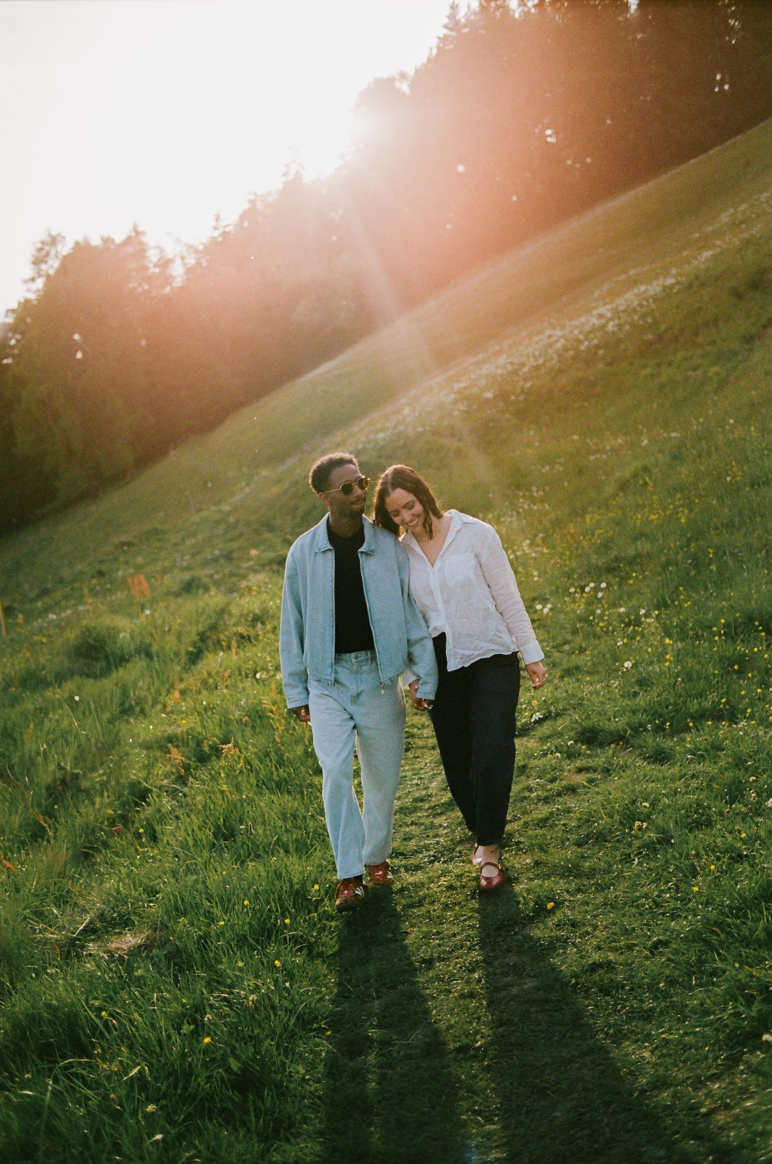 Couple holding hands walking on a sunny green hillside at golden hour with warm sunlight glowing behind them.