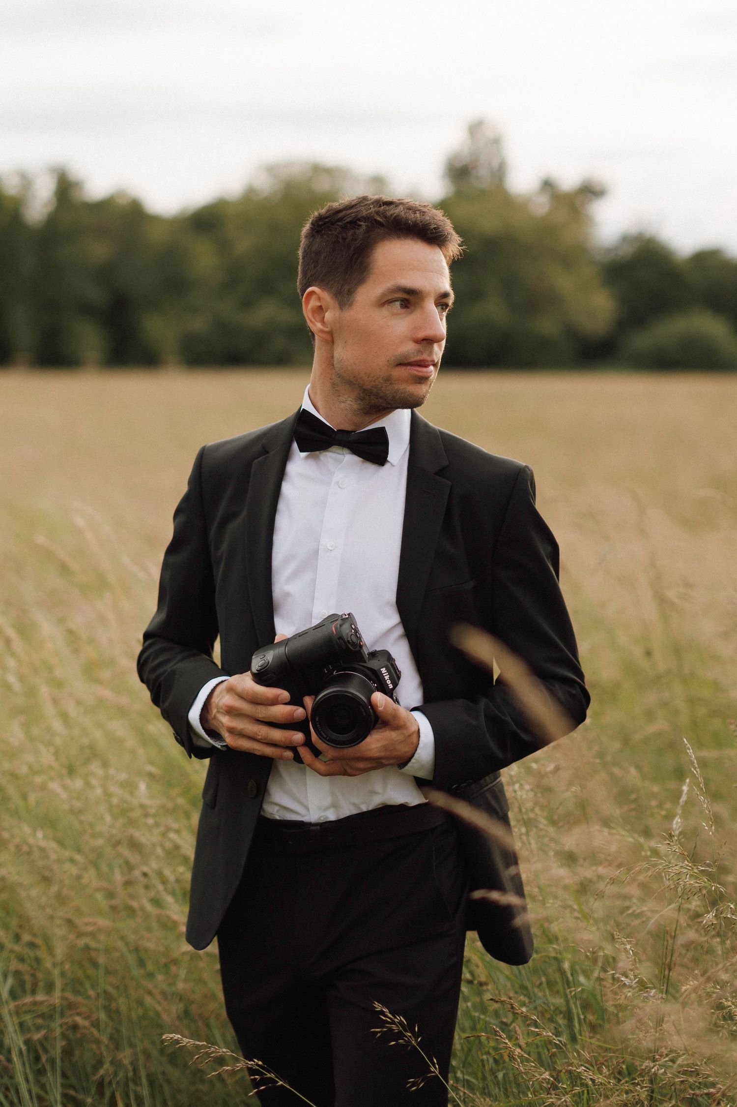 Photographer in a black tuxedo and bowtie holding a camera while standing in a grassy field at golden hour.