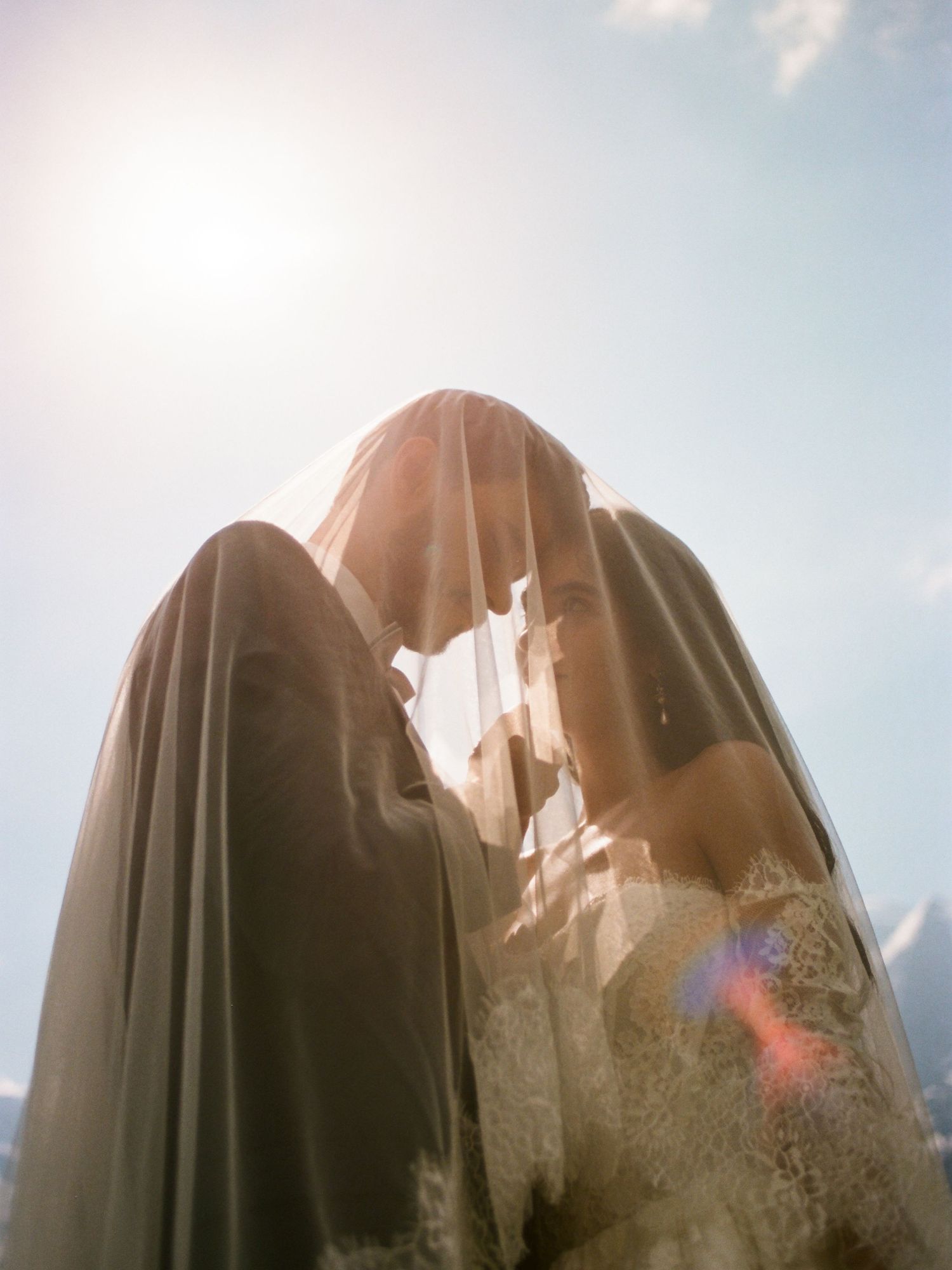Romantic wedding couple sharing an intimate moment under a veil with sunlight and blue sky backdrop.