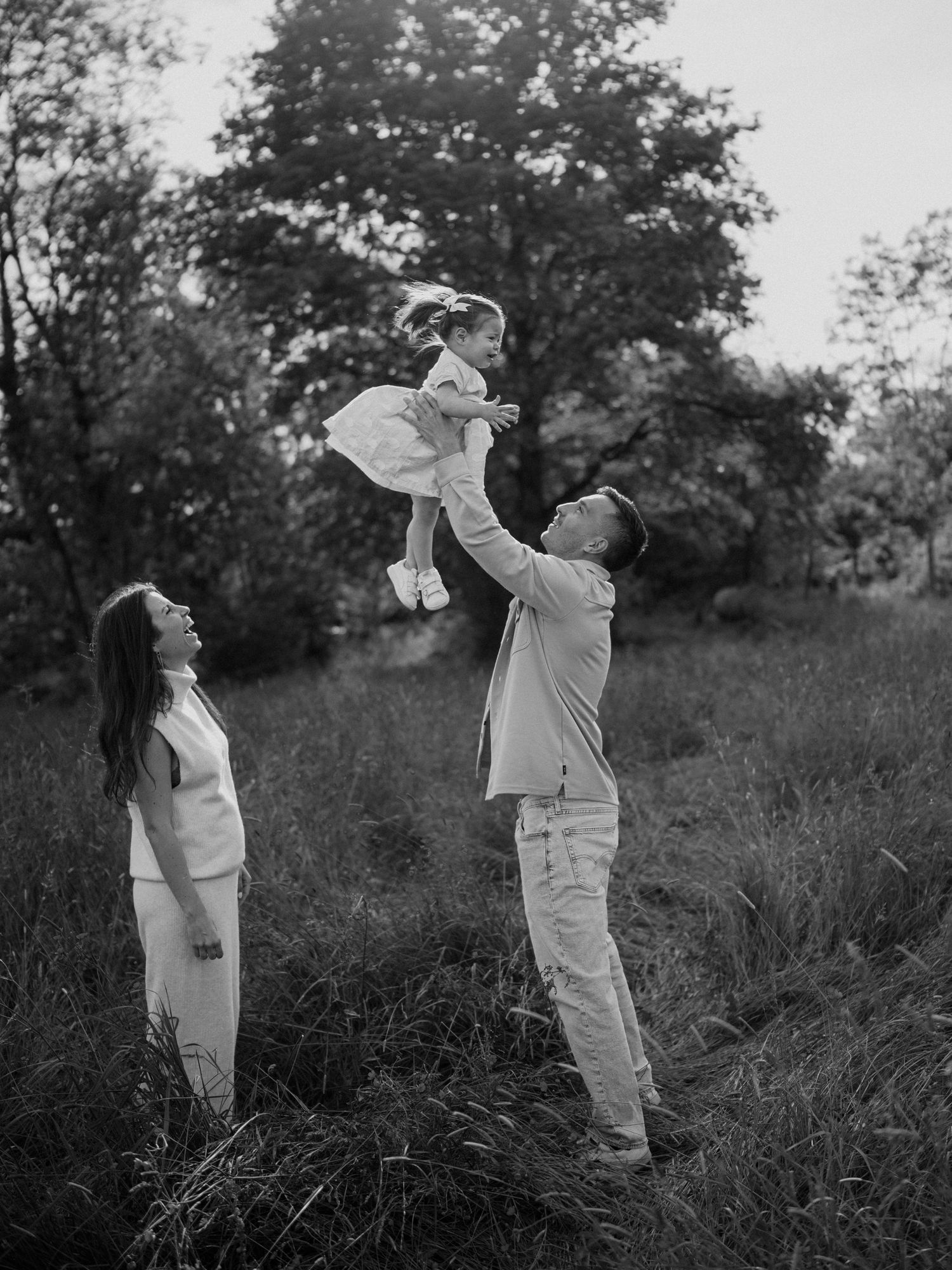 A black and white photo shows a family moment outdoors with parents lifting a small child in a dress against a natural backdrop.