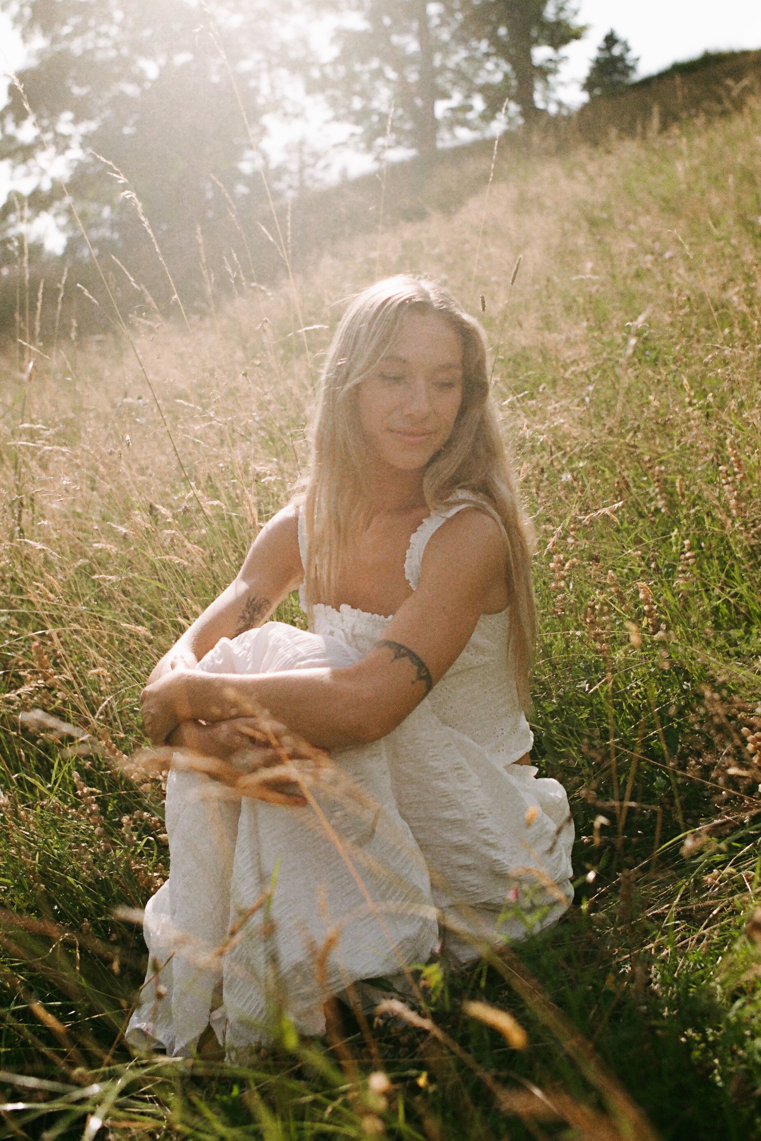 Young blonde woman in white dress sitting in sunlit meadow grass, golden hour light, serene outdoor portrait.