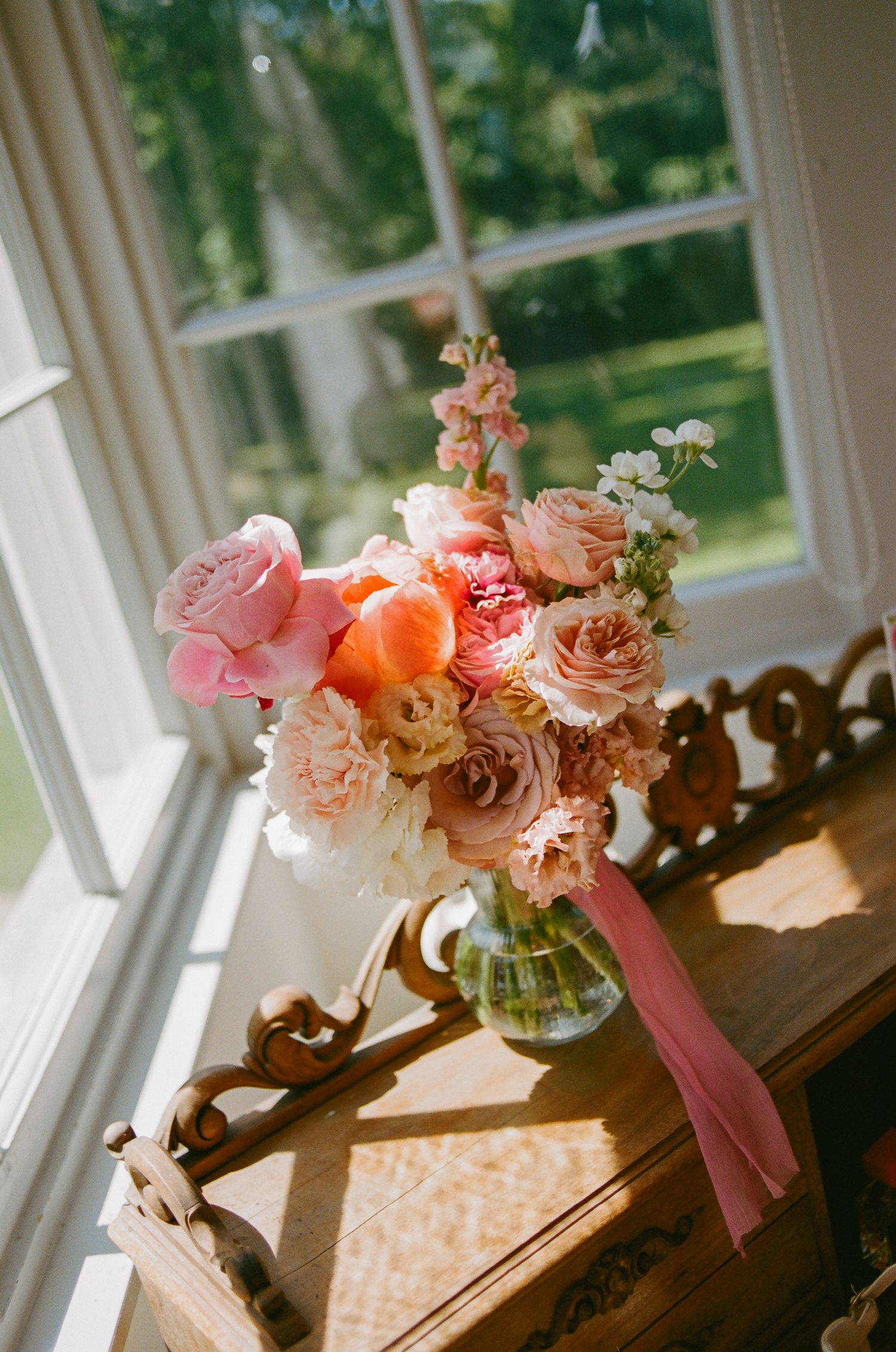 Pink and peach floral bouquet in glass vase with ribbon on ornate wooden desk by sunlit window.
