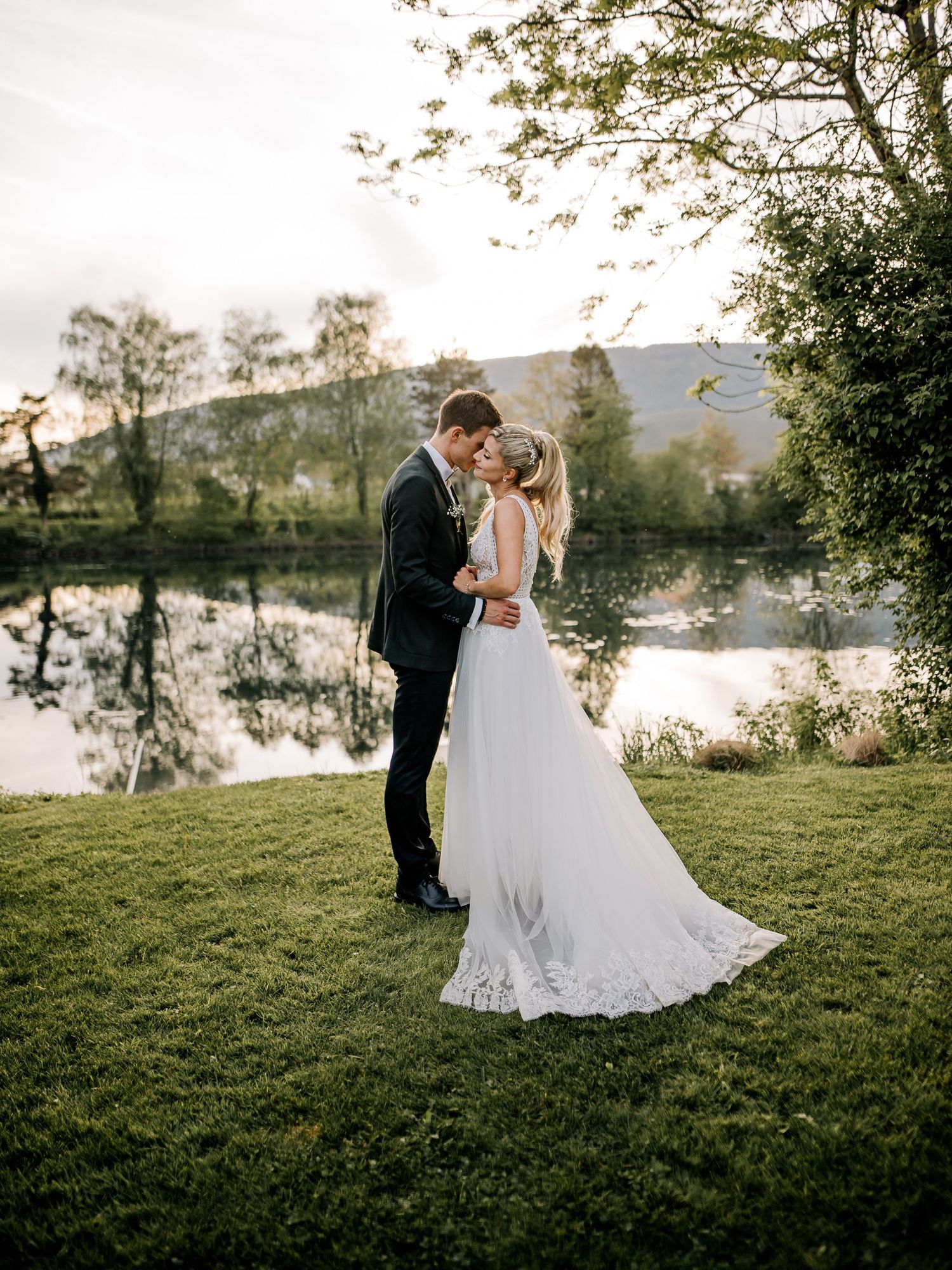 A couple shares romantic wedding photos by a serene lakeside at sunset.