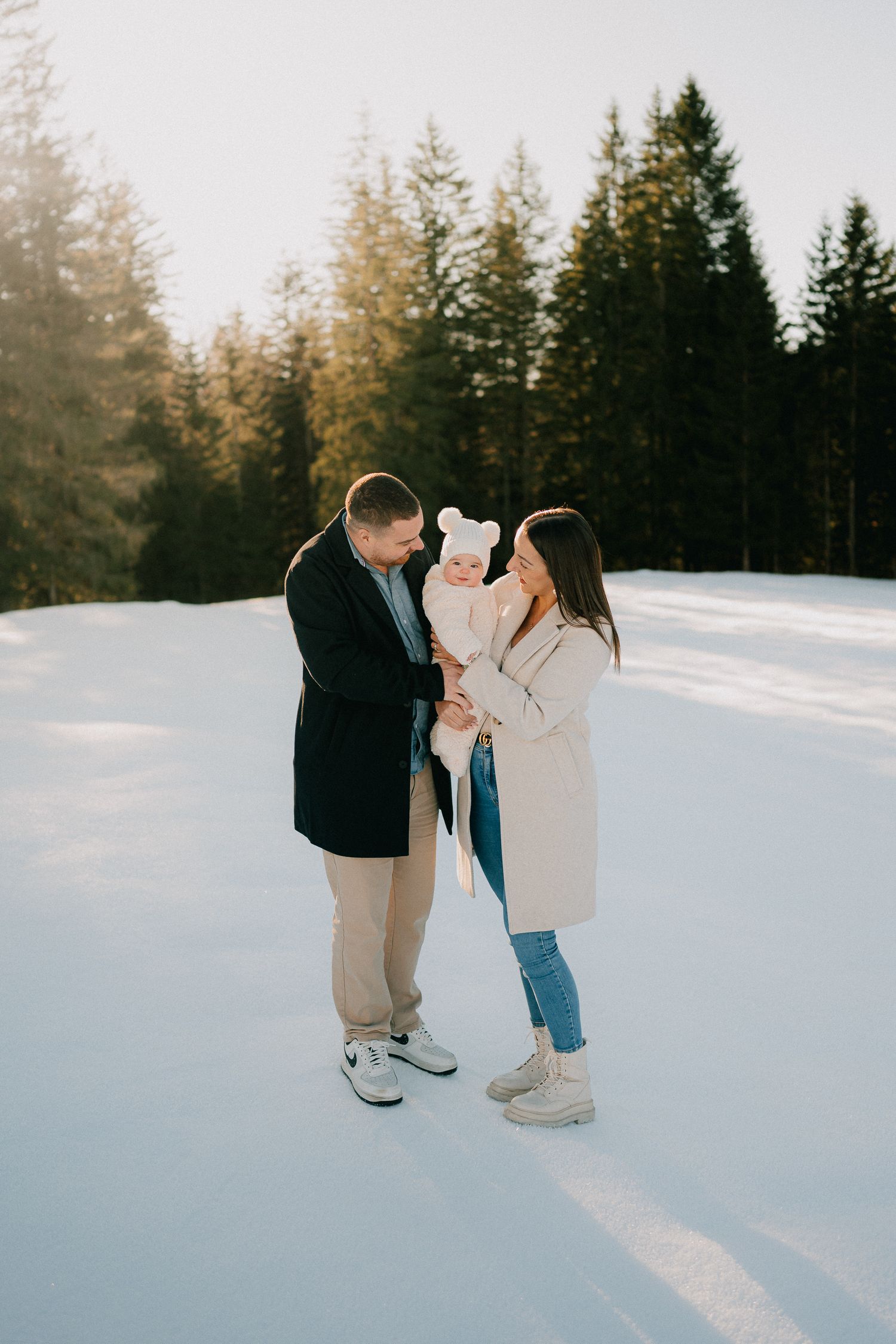 A couple shares an intimate moment in the snow with evergreen trees in the background during golden hour.