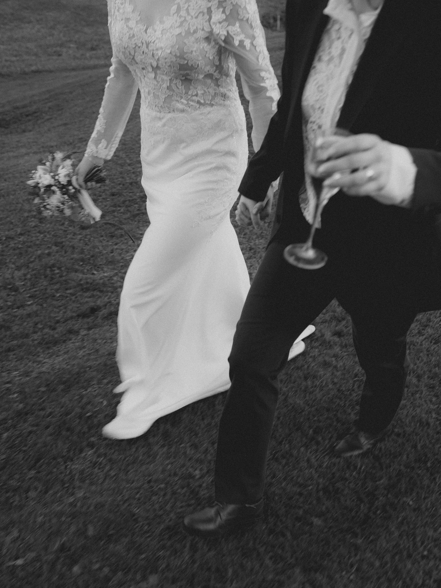 Close-up detail of a white lace wedding dress and black formal suit as couple walks on grass.