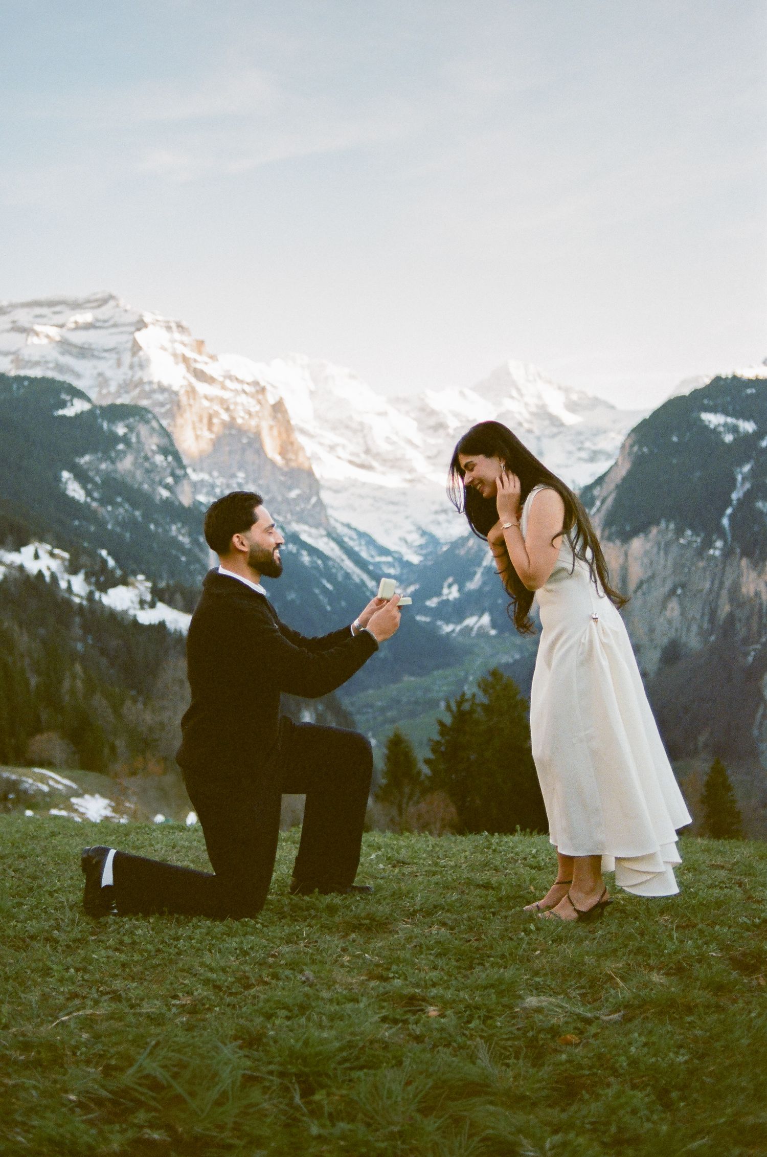 Man proposes on one knee to woman in white dress with stunning Swiss Alps mountains in background.
