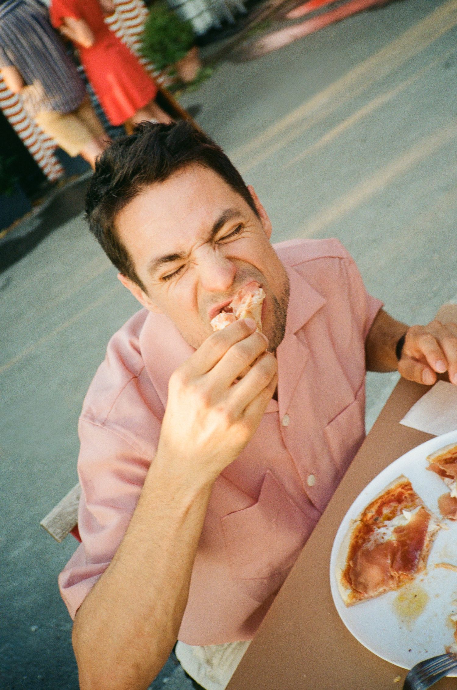 A man in a pink shirt eating a slice of pepperoni pizza outdoors, squinting with enjoyment.