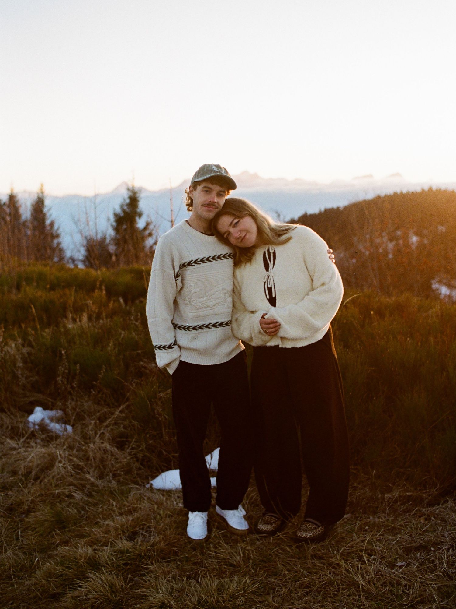 Couple in matching cream sweaters embracing at golden hour on a mountain hilltop with snowy peaks in background.