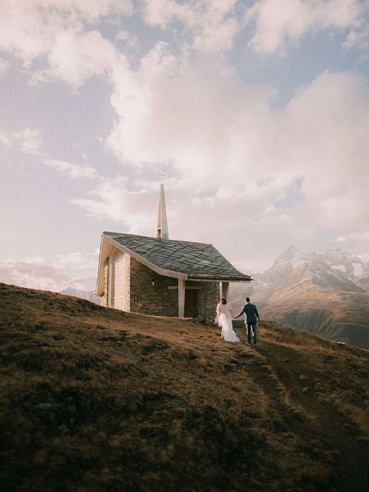Small mountain chapel nestled on a hillside with dramatic alpine peaks in the distance.
