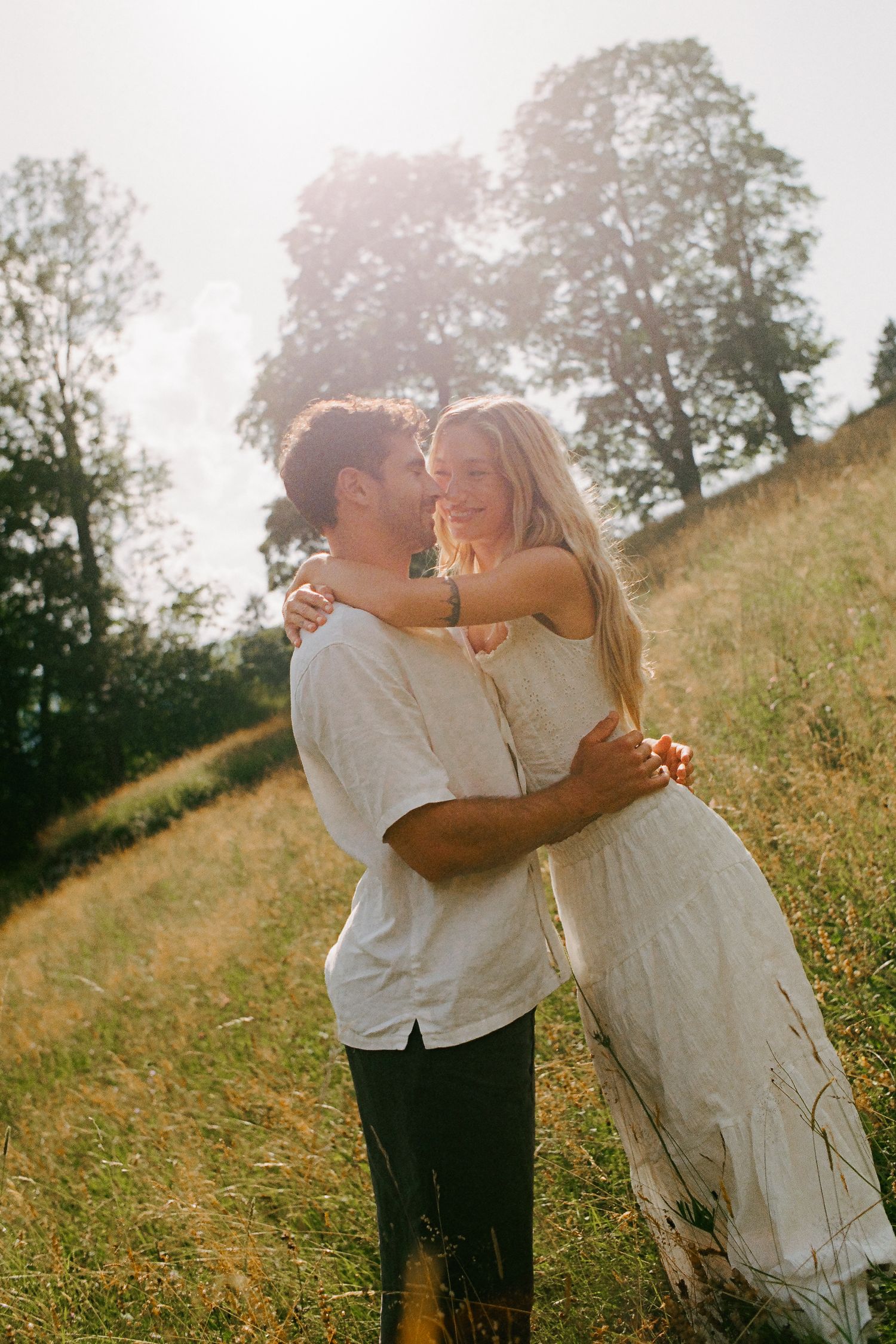 Couple embracing in a sunlit meadow, golden hour light glowing behind trees, woman in white dress smiling.