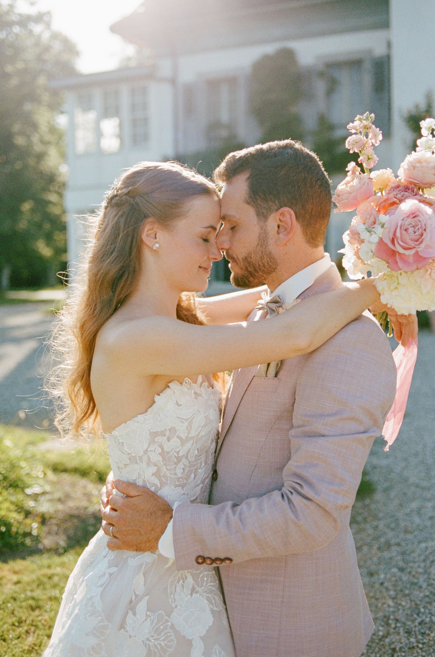 Bride and groom in romantic forehead touch pose, she in lace gown, he in blush suit, holding pink floral bouquet outdoors.