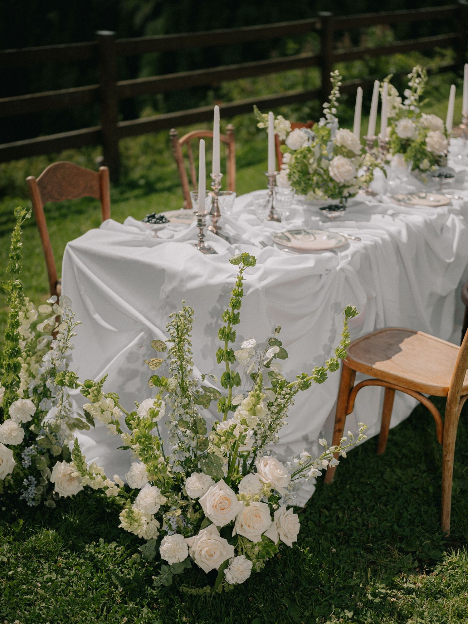 Elegant outdoor wedding table setting with white linens, taper candles and cascading white floral arrangements on grass.
