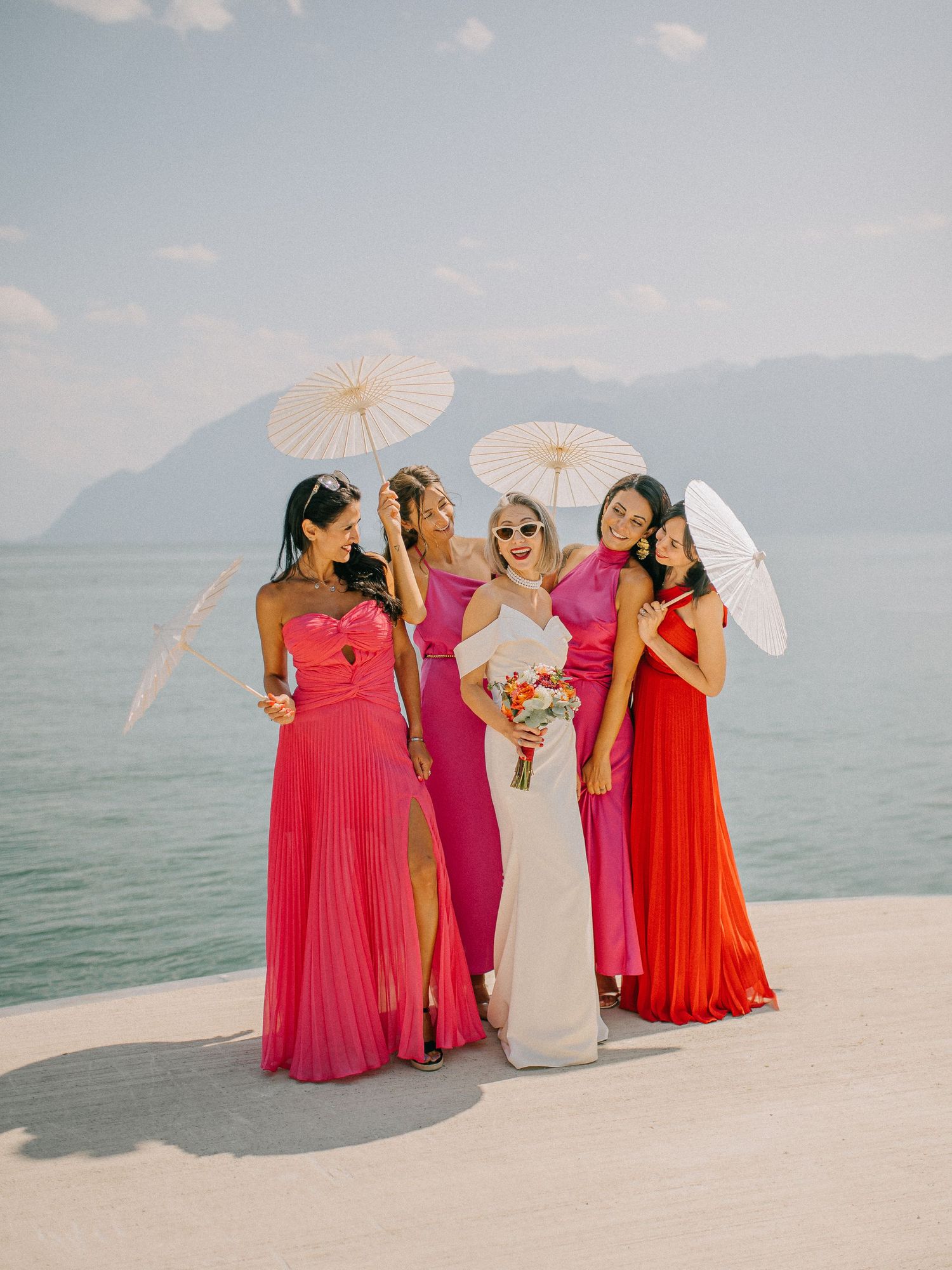 Bridal party in vibrant pink and red dresses posing with parasols by lakeside.