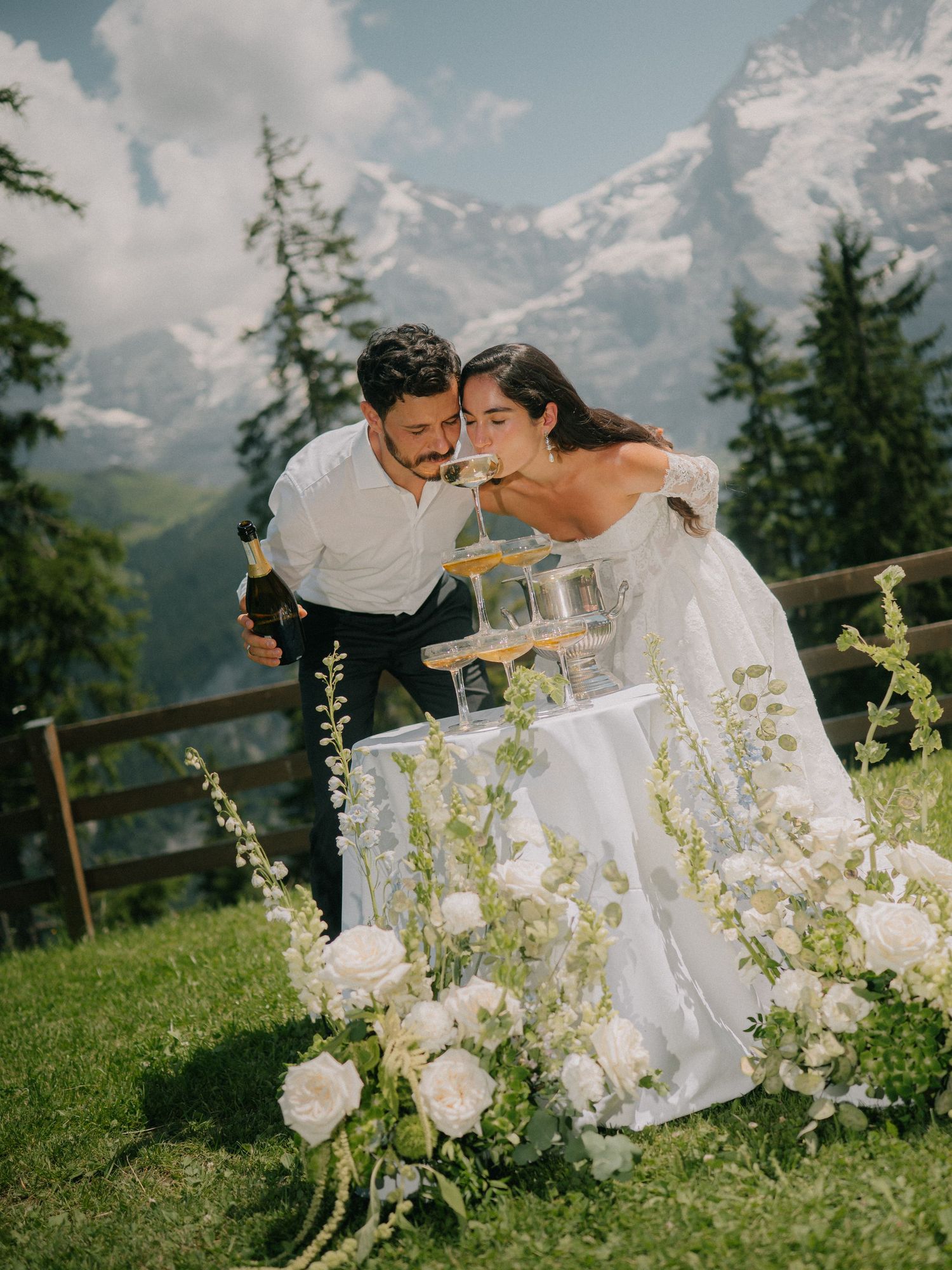 A romantic couple shares an intimate moment on a grassy hillside with snow-capped mountains and white flowers in the background.