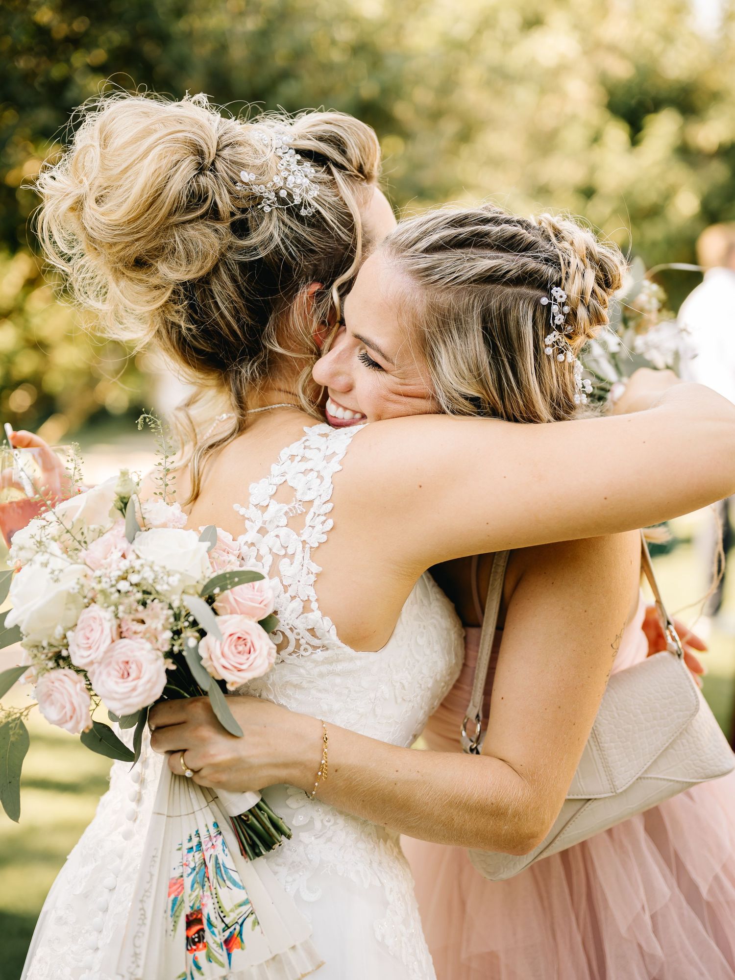 Bridesmaids in pink dresses embrace and celebrate with bouquets during an outdoor wedding ceremony.