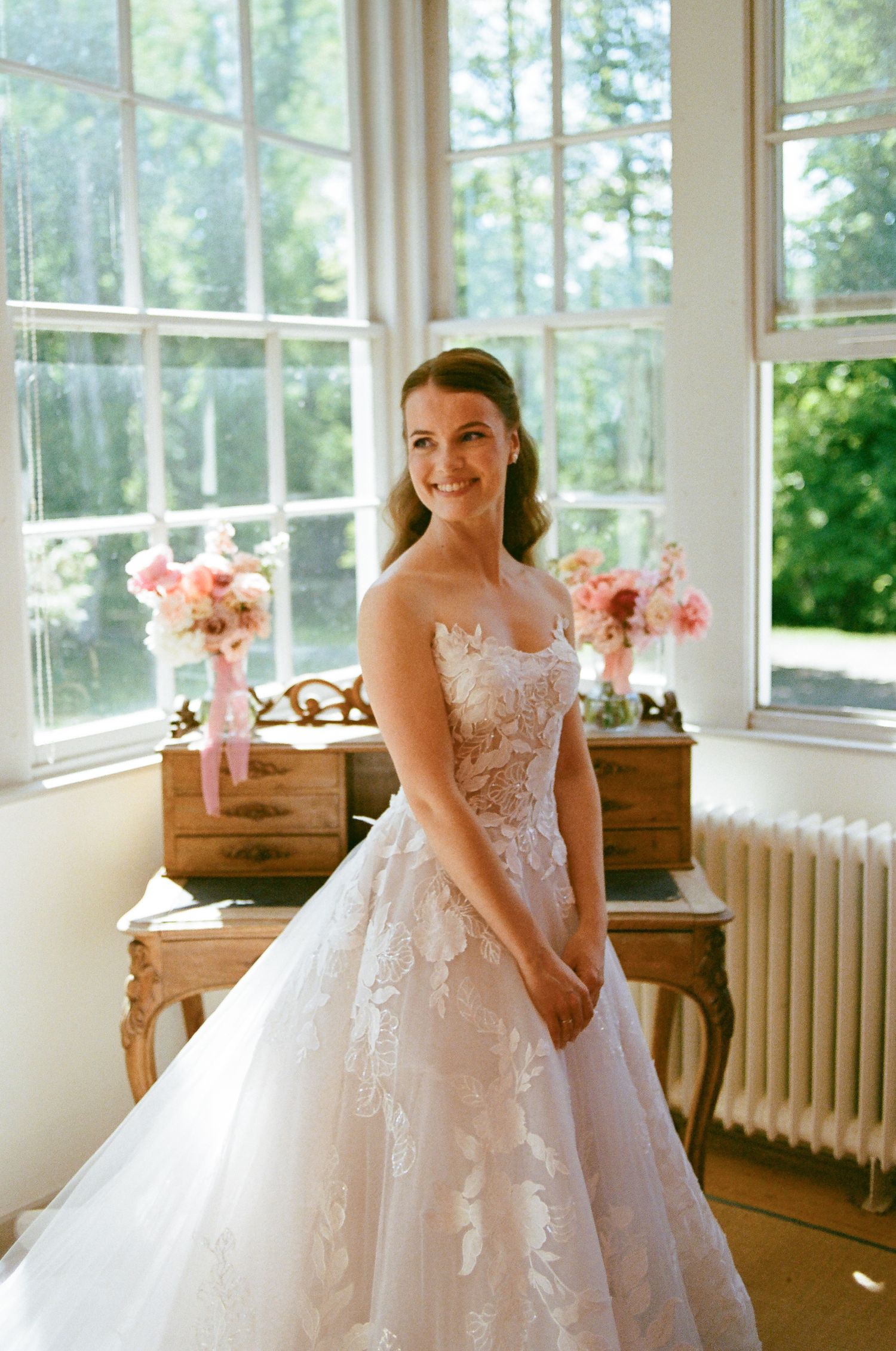 Smiling bride in strapless lace ball gown wedding dress stands before sunlit windows with pink floral arrangements.