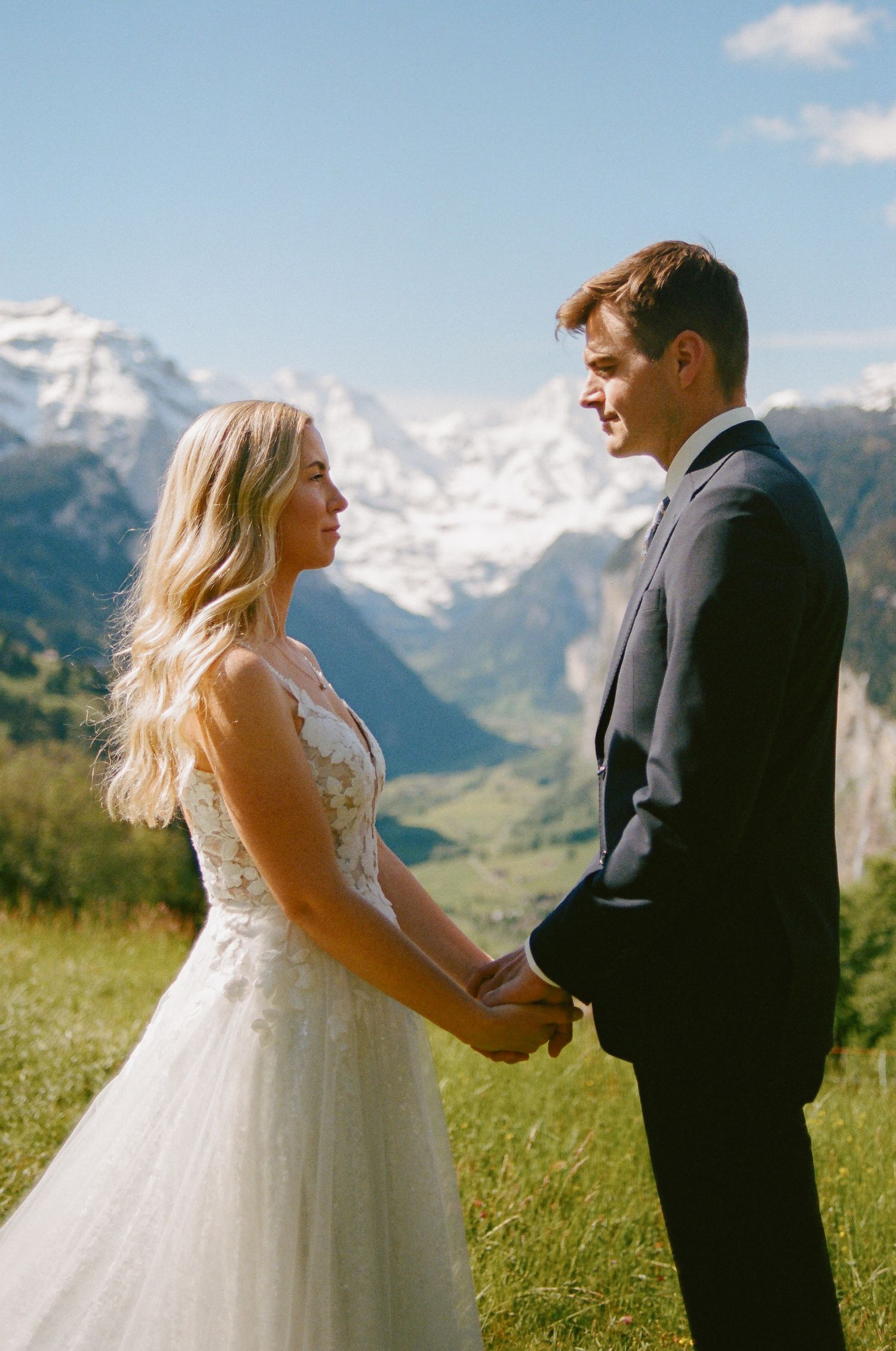 Bride and groom hold hands in Swiss Alps meadow with snow-capped mountains and valley backdrop on sunny wedding day.