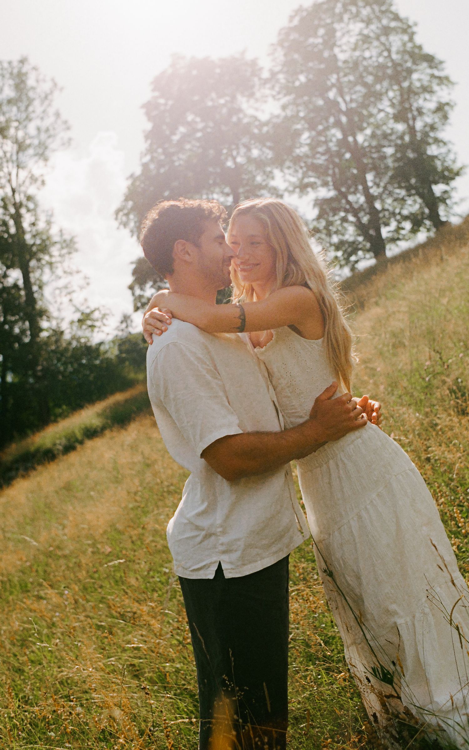 A couple embraces and almost kisses in a sunlit meadow, the woman in a white dress and man in a white shirt.