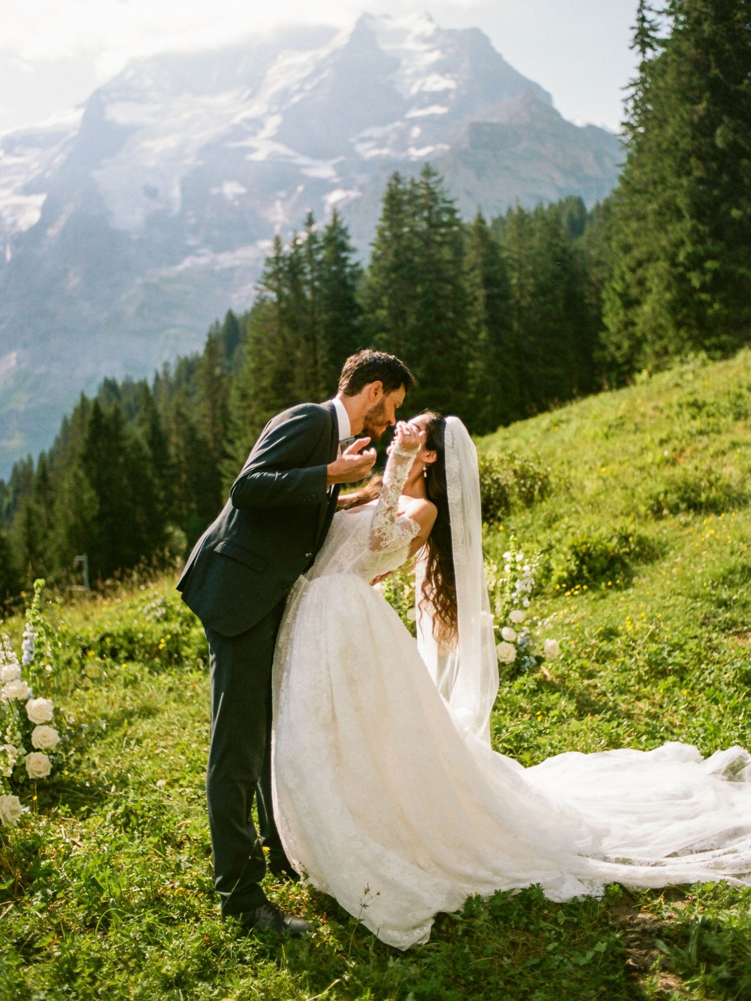 Bride and groom share a romantic kiss on a lush green alpine meadow with snow-capped mountains in the background.