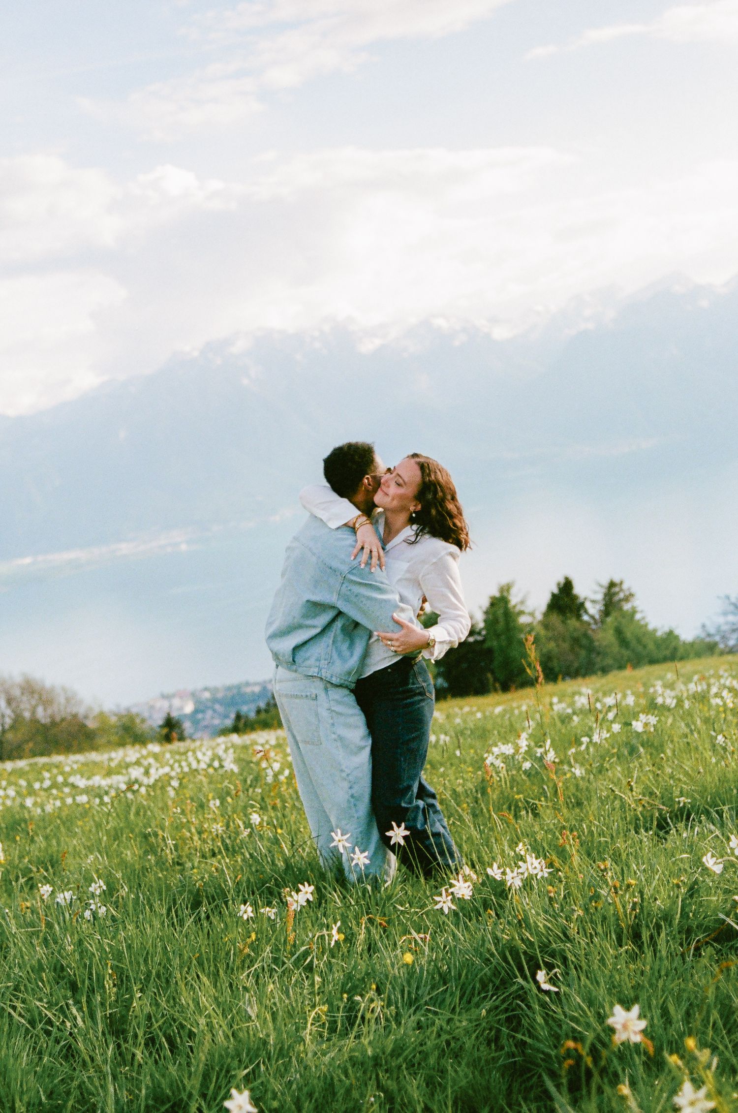 Couple embracing and kissing in a blooming meadow with white flowers, mountains and lake in the background.
