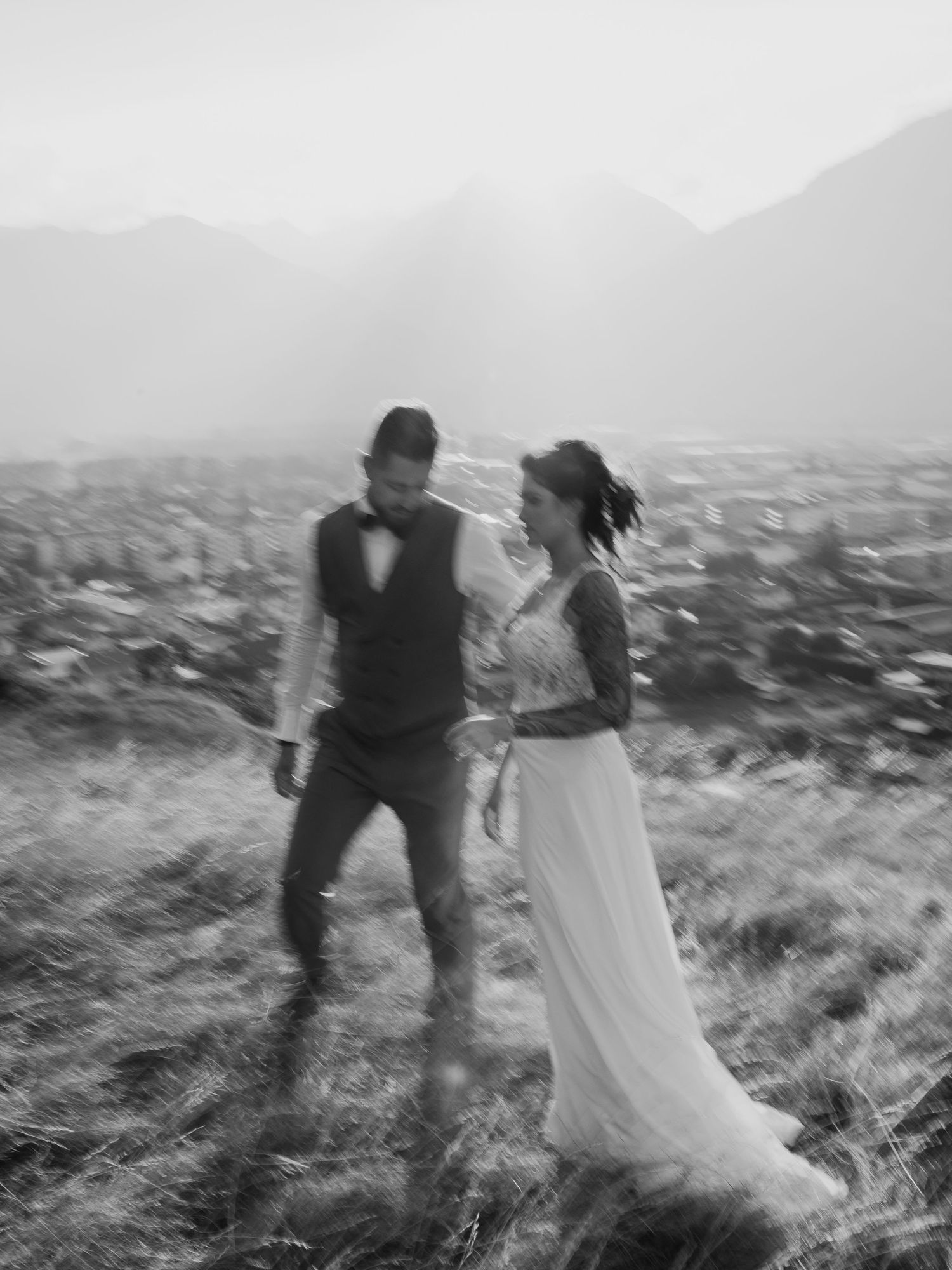 A couple spins playfully in a windswept field with mountains in the distance.