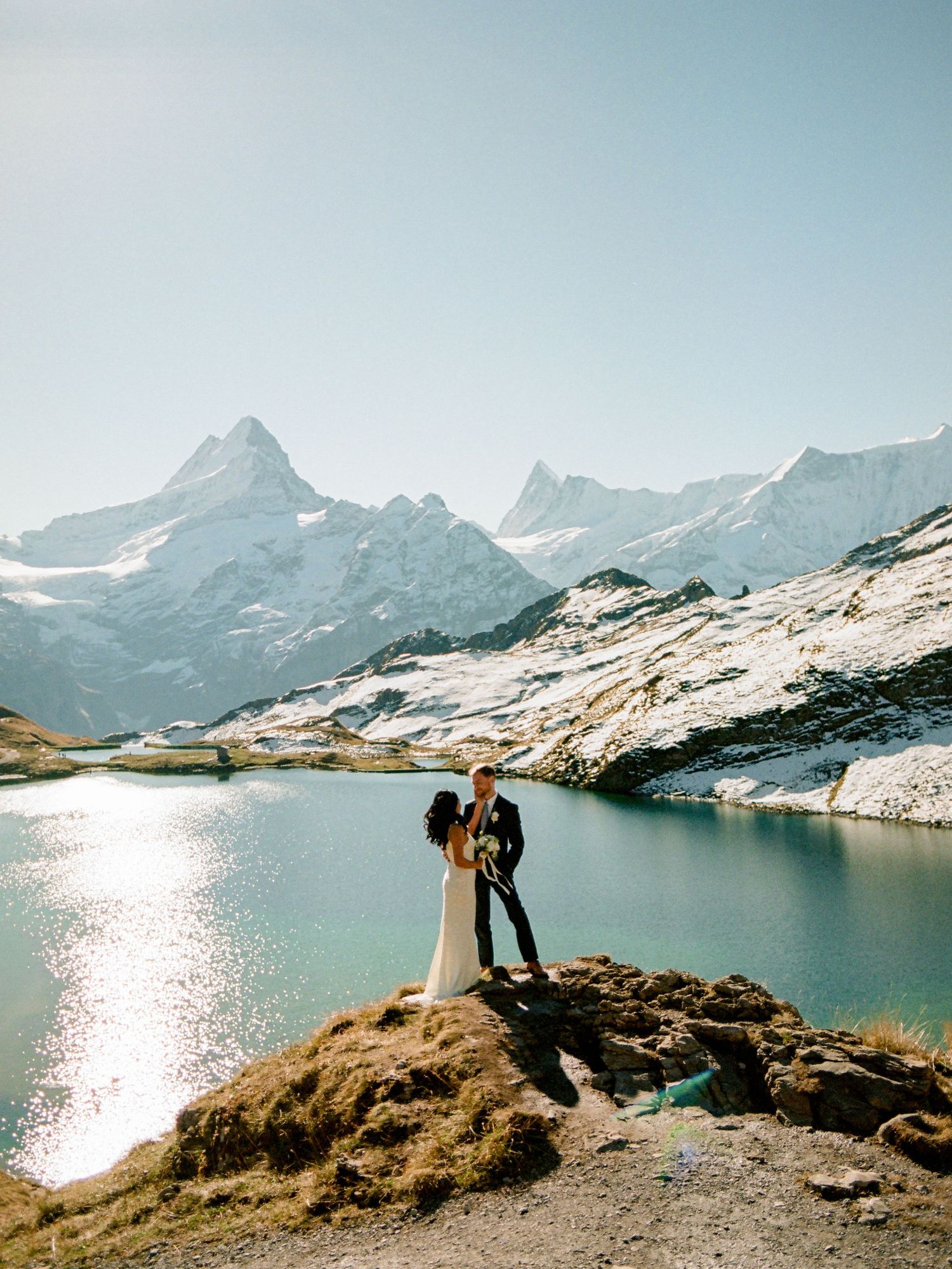 Bride and groom embrace on rocky alpine shore with snow-capped Swiss mountains and turquoise lake backdrop.